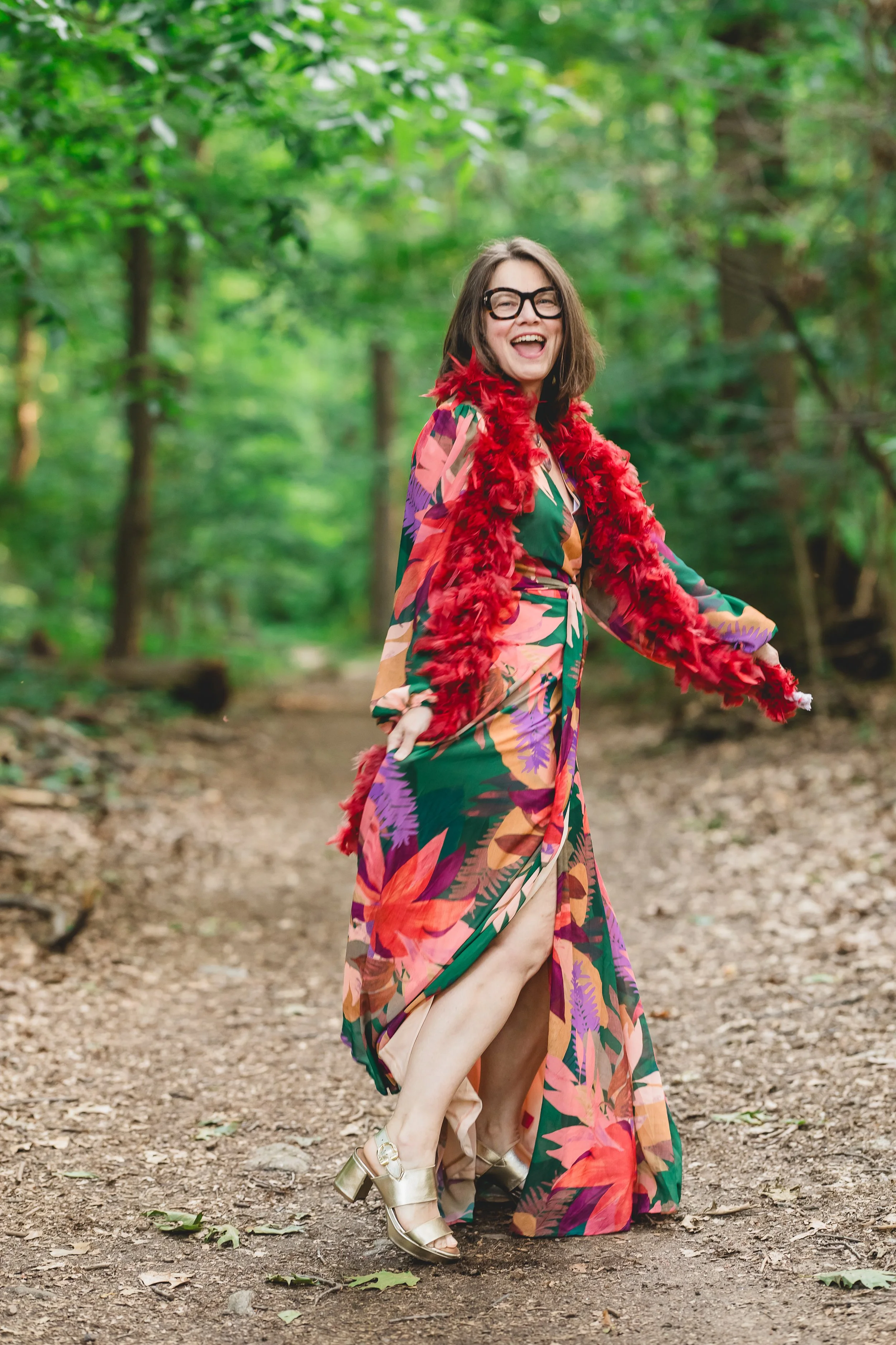 Deb Valentine standing joyfully on a wooded path, wearing a colorful flowing dress, gold heels, glasses, and a red feather boa, smiling and moving playfully among green trees.