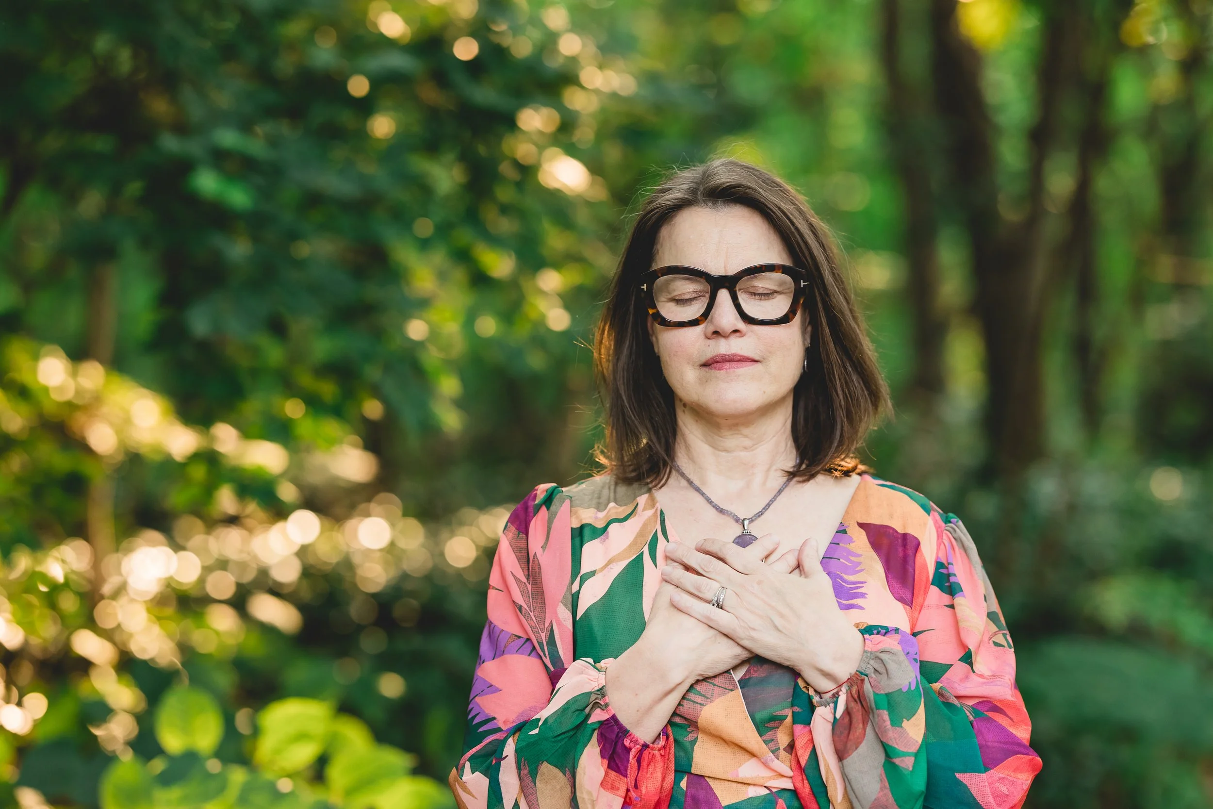 Portrait of white woman with brown hair  wearing glasses and a colorful patterned top, standing outside in natural light surrounded by green leaves, with her eyes closed and both hands gently placed over her heart.