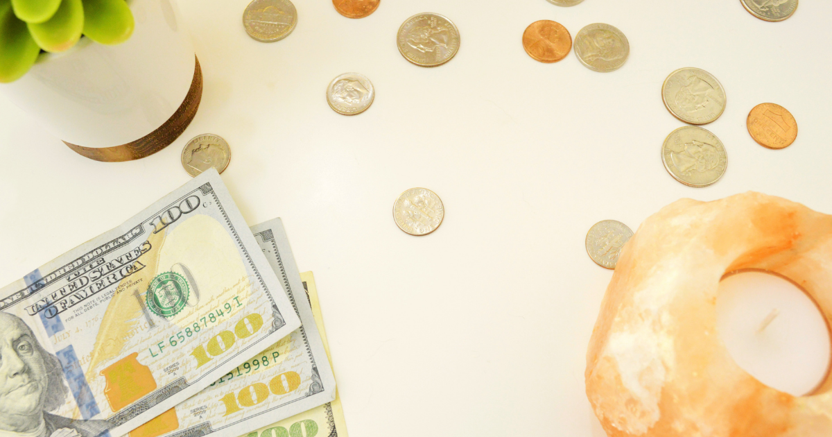 Flat lay of US dollar bills, coins, a small potted succulent, and an orange Himalayan salt candle holder on a light cream surface.