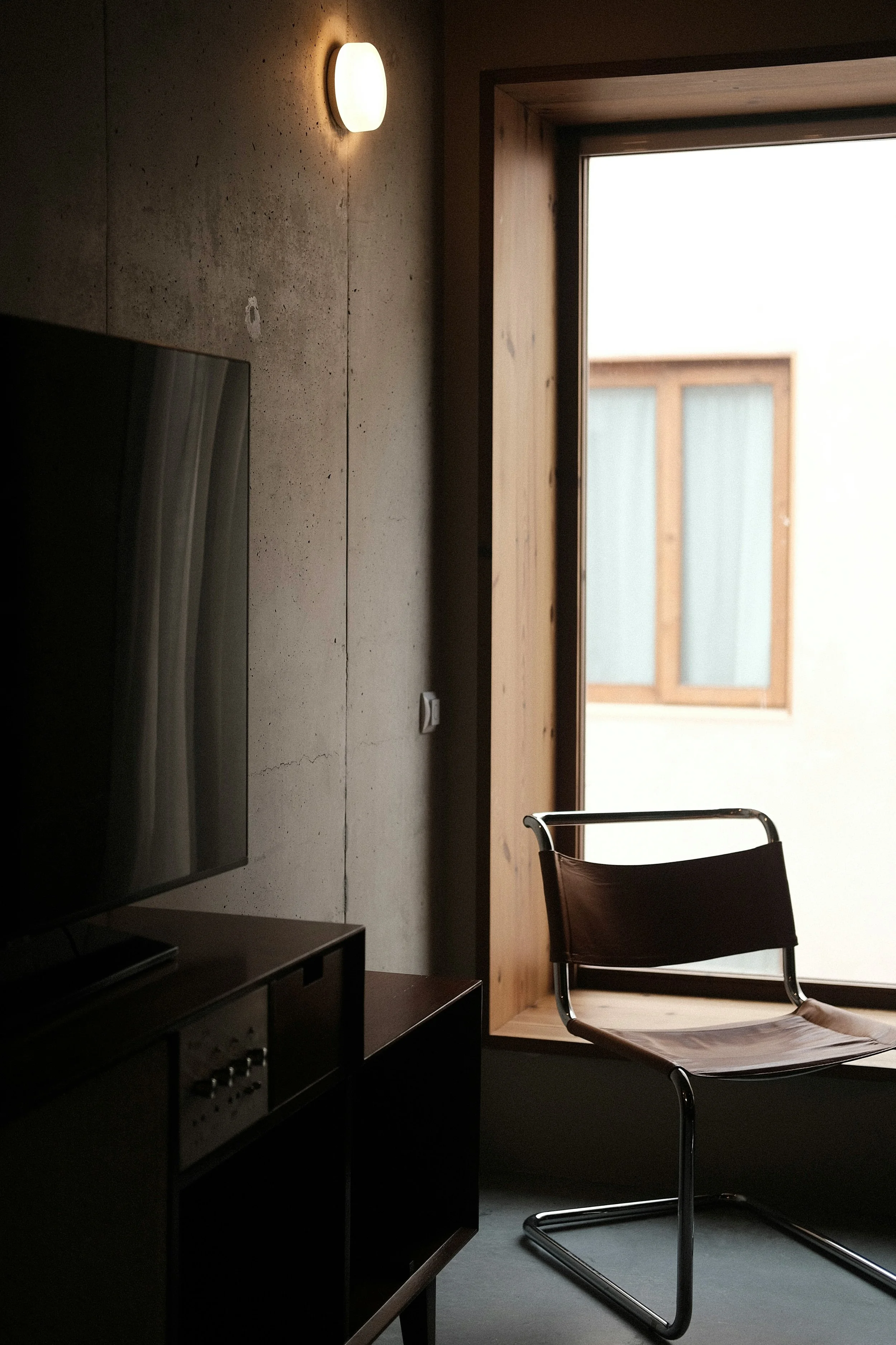  Interior of a modern room with a concrete wall, a mounted circular light, a flat-screen television on a black stand, a wooden window frame, and a black chair near the window.