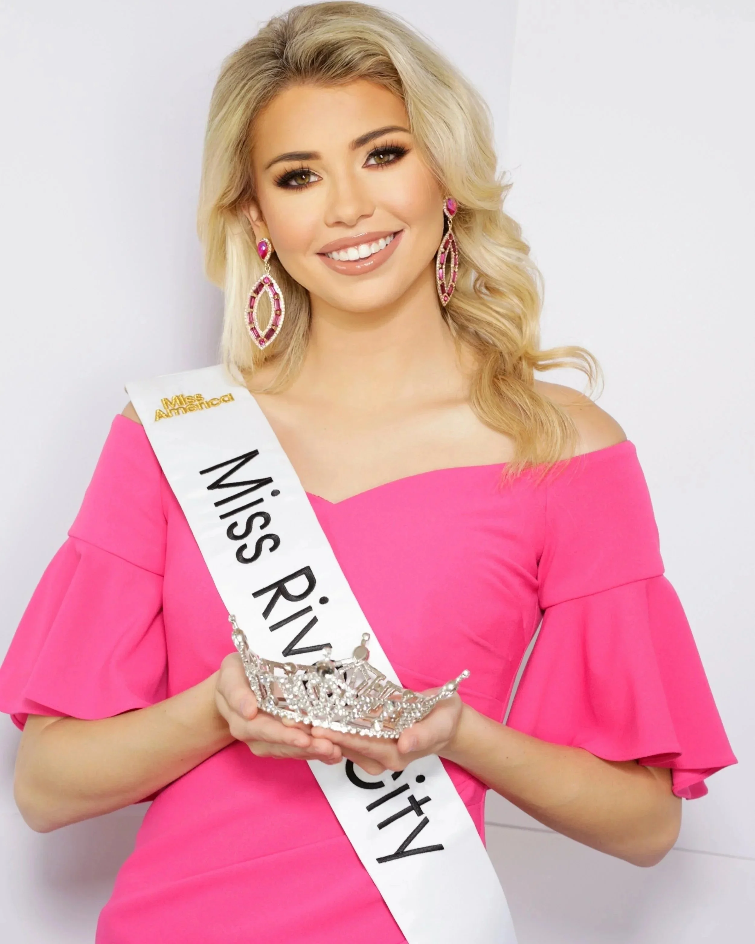 A woman with blonde hair wearing a pink dress and large pink earrings, holding a crown, and wearing a sash that reads 'Miss RIV Miss America'.