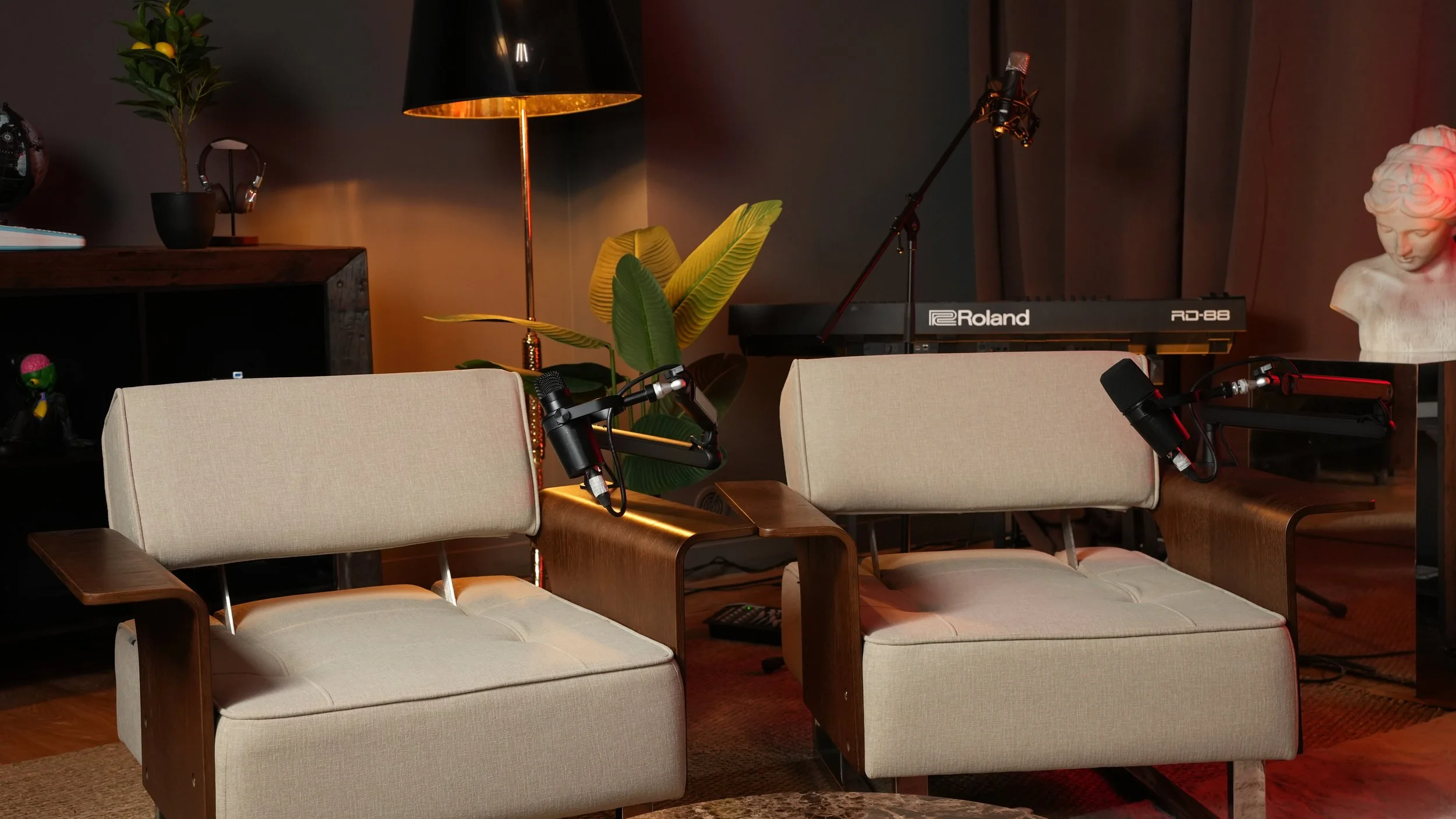 Un homme assis sur un fauteuil blanc, parlant dans un microphone, avec un fond lumineux orange et une plante décorative.