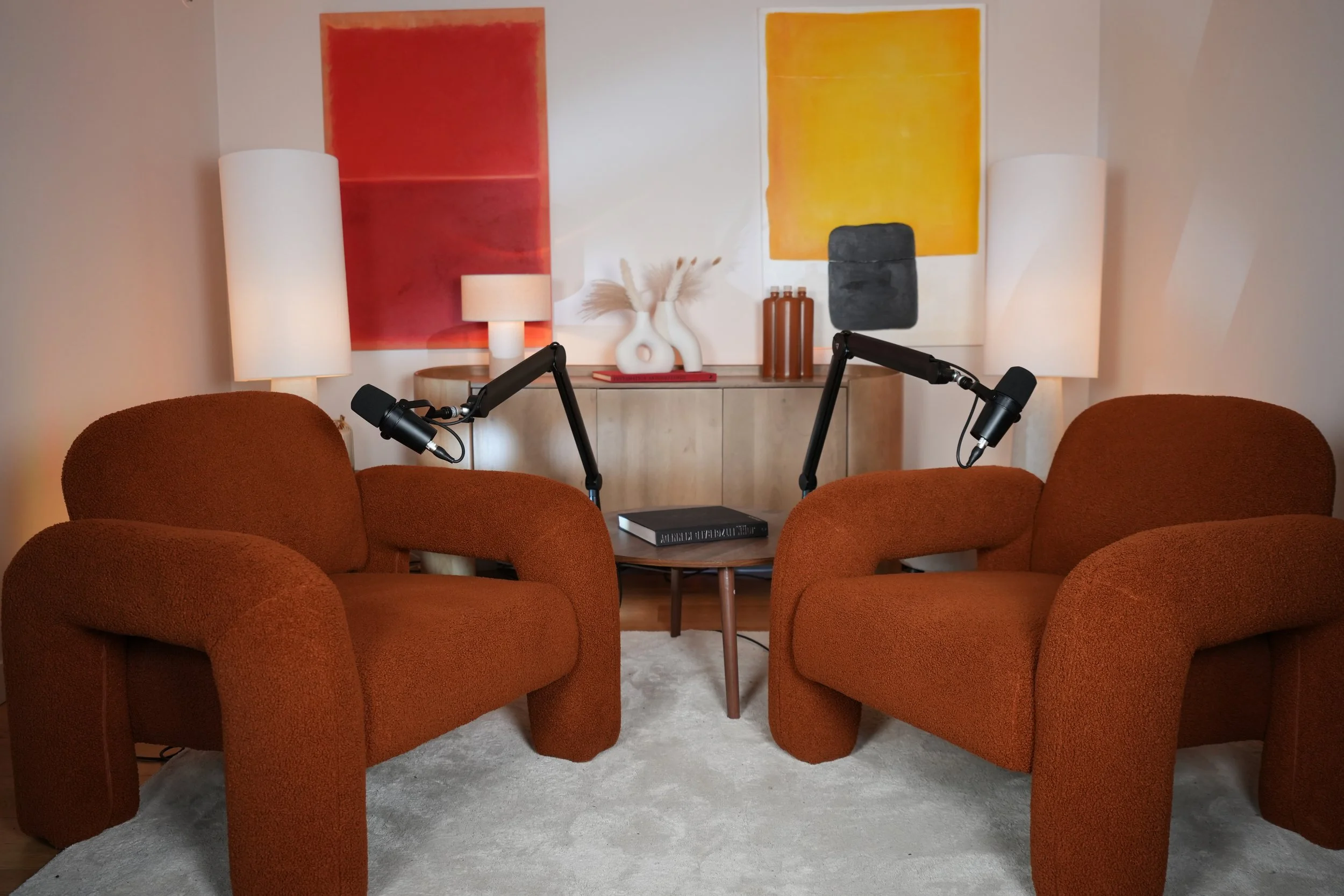 Un homme assis dans un studio, tenant un téléphone et portant une casquette, un pull avec un logo, et assis devant un microphone, avec un arrière-plan sombre et des décorations végétales.