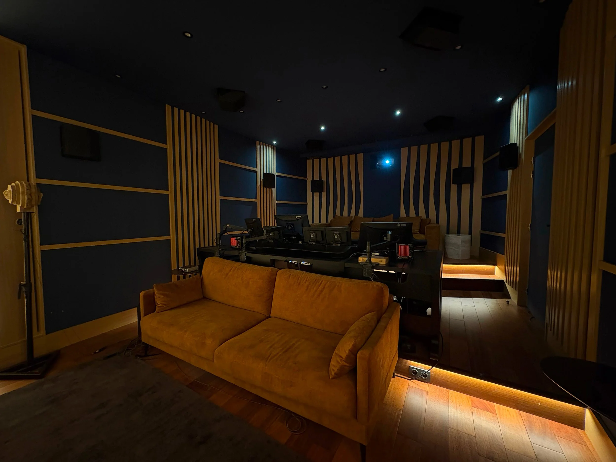 Salle de contrôle avec un canapé orange, plusieurs écrans et équipements audiovisuels, éclairage tamisé.