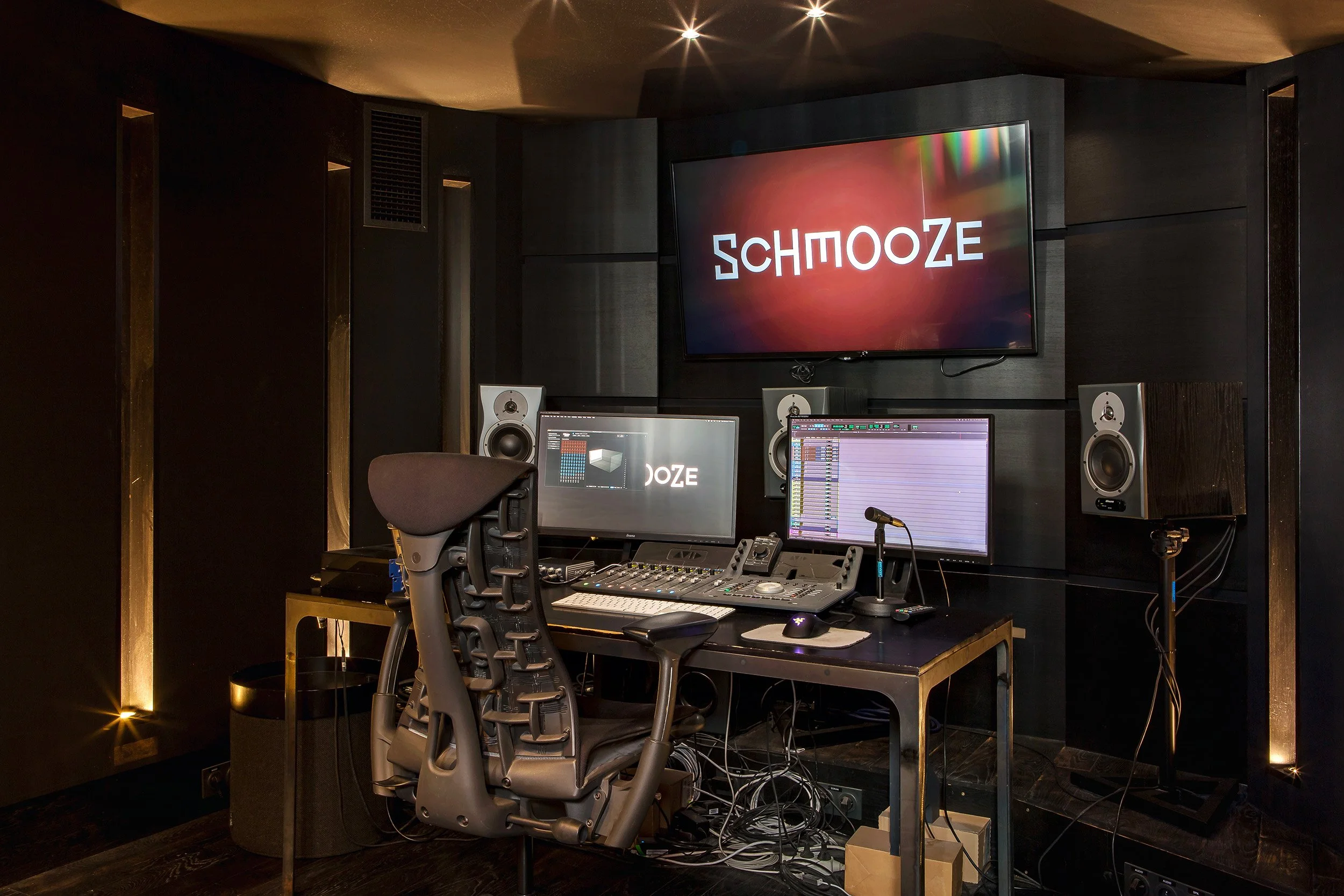 Studio d'enregistrement avec deux écrans, haut-parleurs et équipements audio, mur noir avec grand écran affichant 'SCHMOOSE'