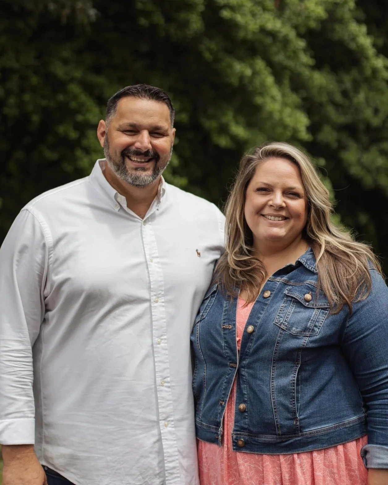 A smiling couple standing outdoors with green trees in the background. The man is wearing a white button-up shirt and jeans, and the woman is wearing a denim jacket over a pink dress with a smartwatch on her wrist.