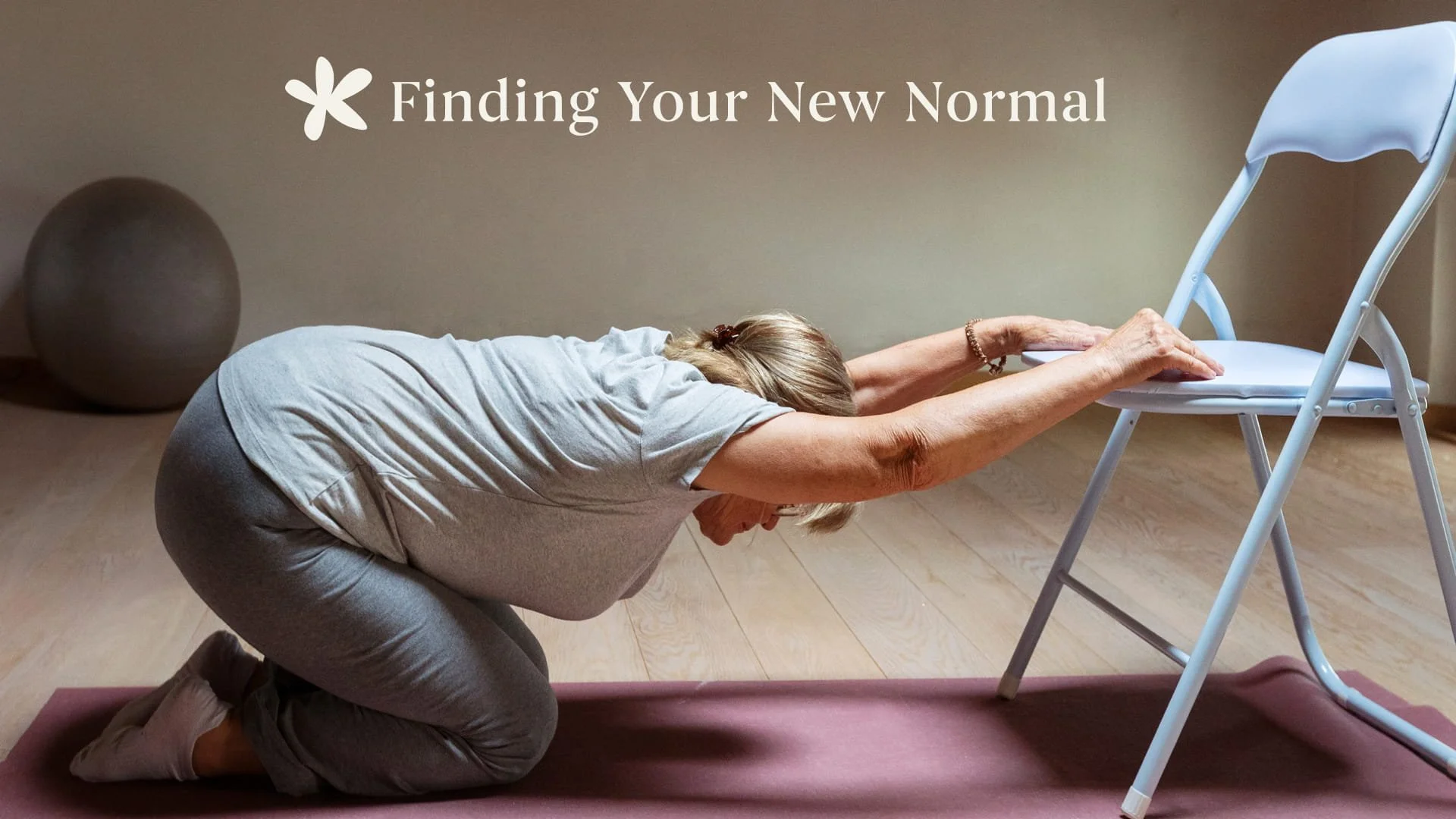 A woman kneeling in child's pose on a yoga mat using a chair to support her arms