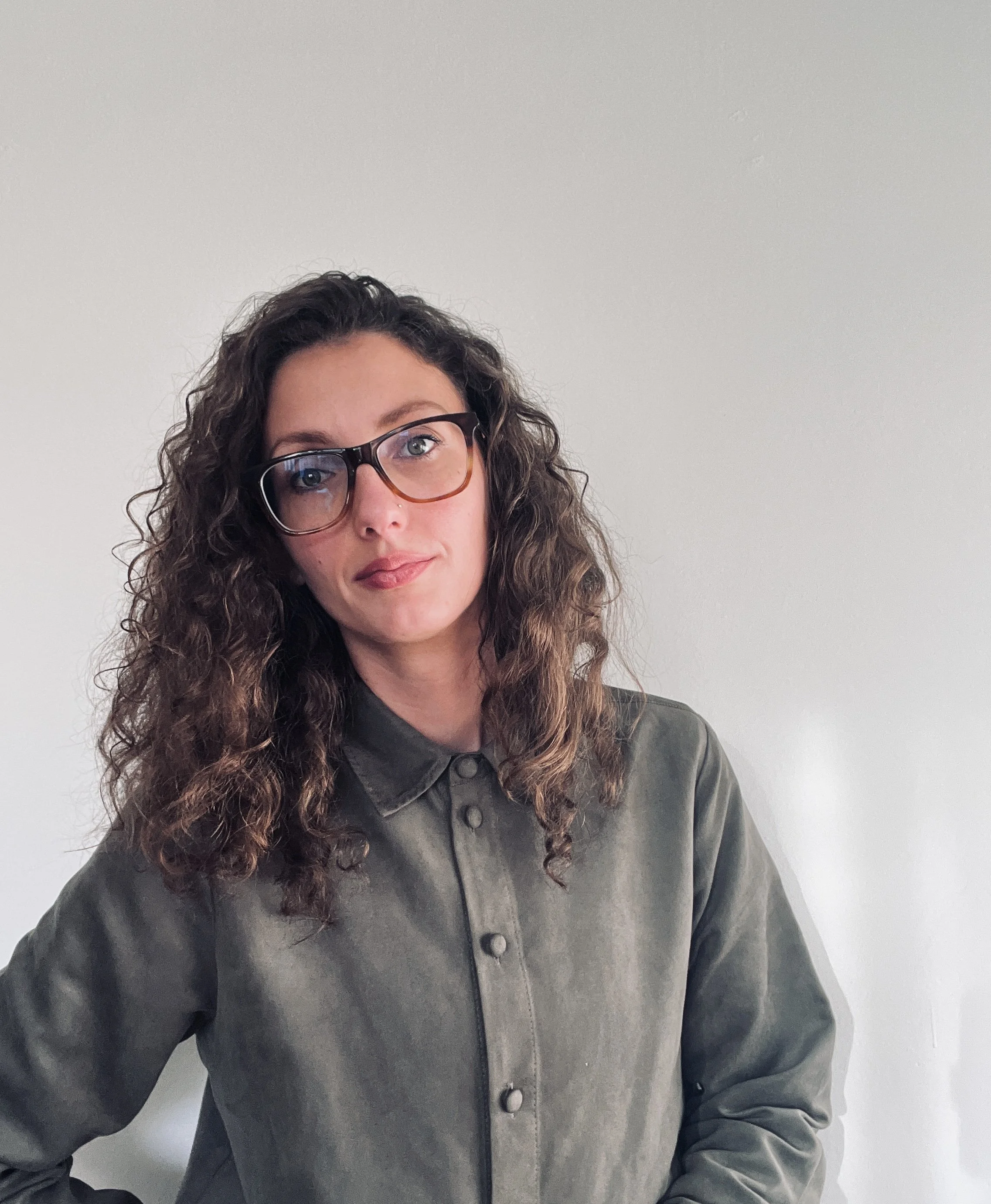 Lauren has shoulder length brown curly hair, green eyes and brown glasses. She is standing in front of a blank white wall and wearing an olive green button down shirt.