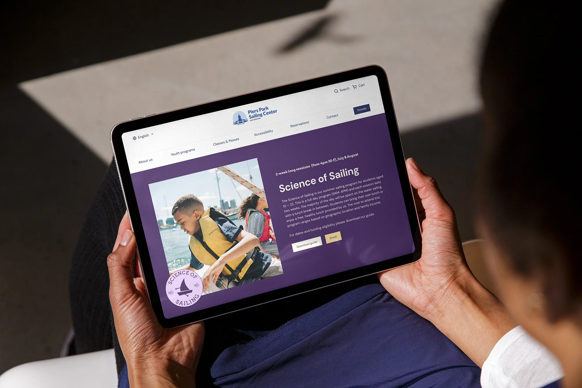 An ipad screen with one of the sailing program service pages 'science of sailing' showing a young boy in a life jacket leaning over the side of a boat to look at the water