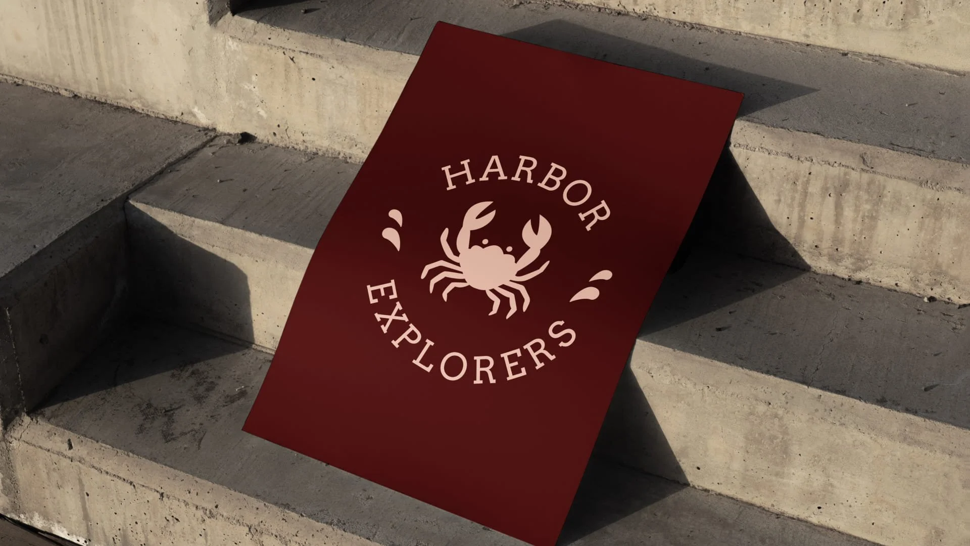Brand poster for Harbor Explorers leaning against concrete steps, featuring a minimalist crab illustration surrounded by curved serif typography on a deep red background.