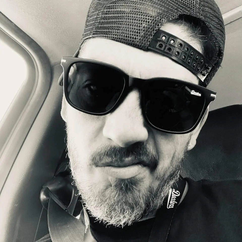 A man with a beard and mustache wearing a backward baseball cap, large sunglasses, and a black shirt, sitting next to an airplane window.