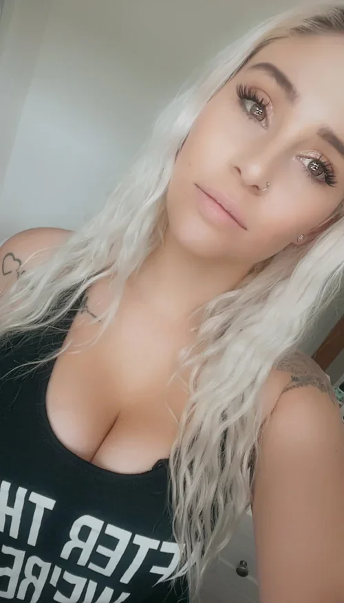A young woman with platinum blonde hair, light gray eyes, and a nose ring taking a selfie indoors. She is wearing a black tank top with white text and has tattoos on her shoulders.