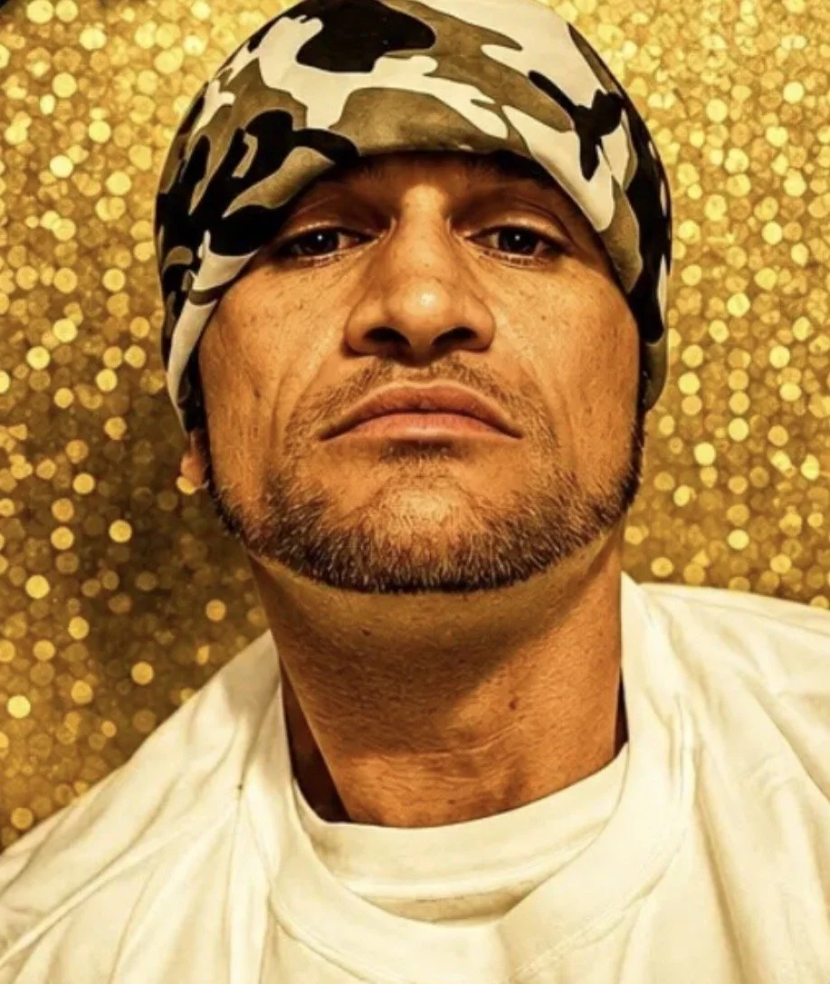 Close-up of a man's face with a serious expression, wearing a camouflage headscarf, against a gold, glittery background.