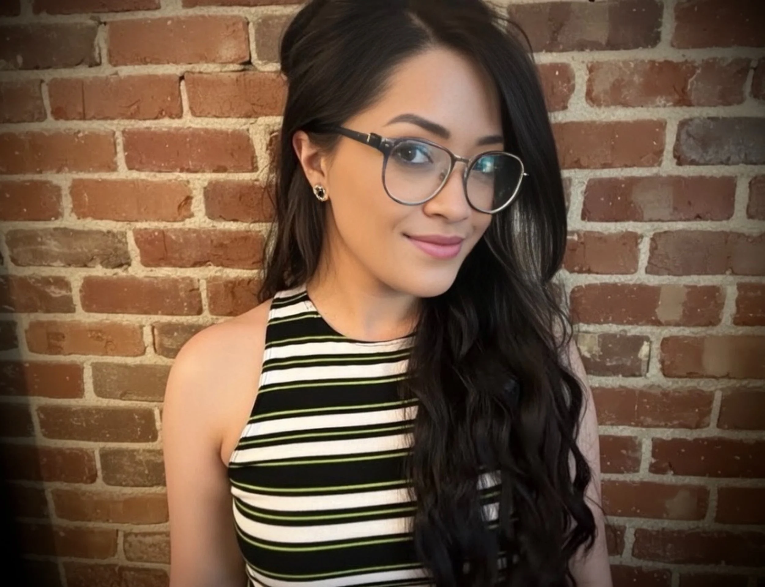 A young woman with long dark wavy hair, wearing glasses and earrings, smiling in front of a brick wall.