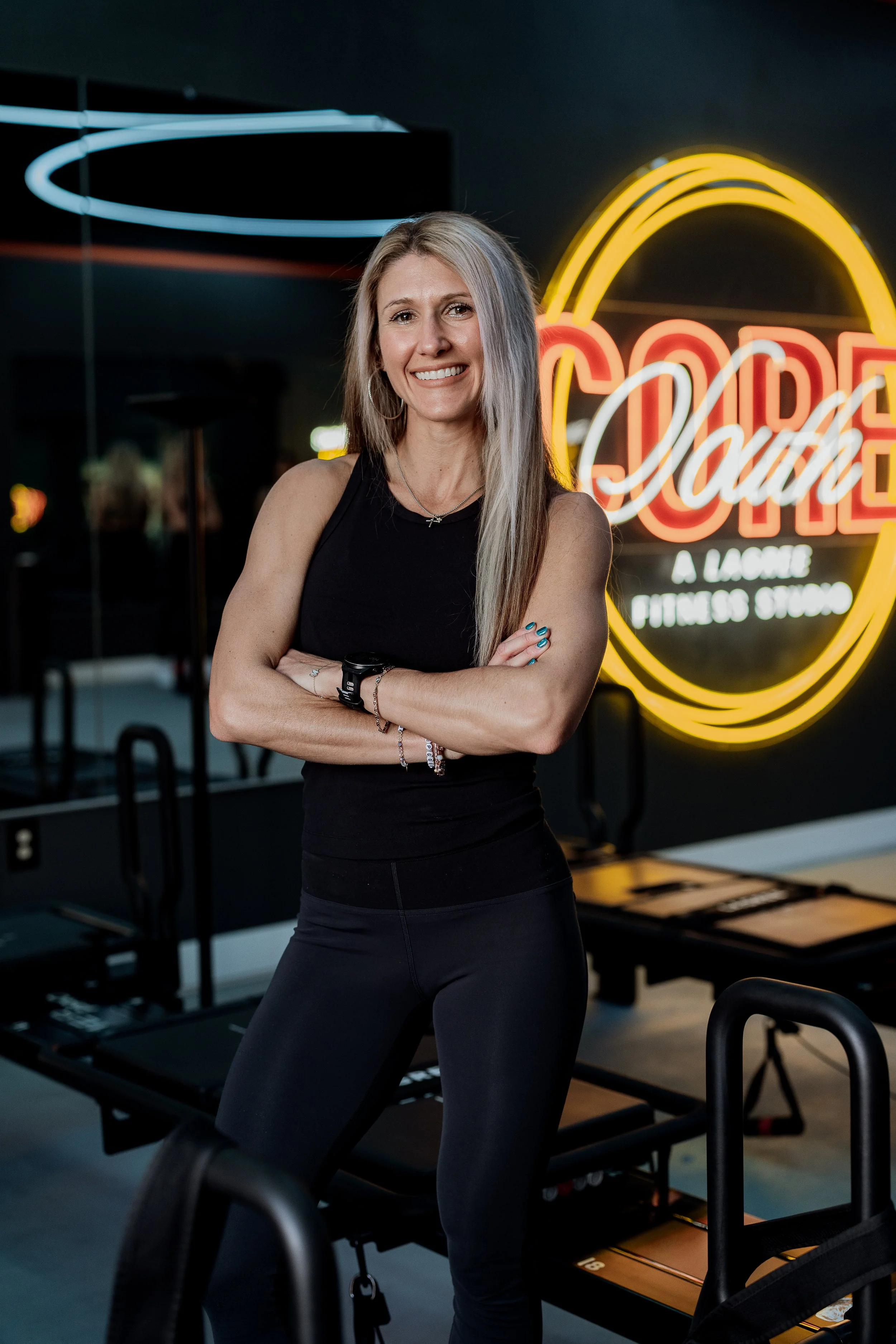 A smiling woman with long blonde hair stands with her arms crossed in front of a neon sign that reads 'Cobra A Lacrosse Fitness Studio.' She is wearing a black sleeveless top and black leggings, inside a fitness studio with exercise equipment including pilates reformers.