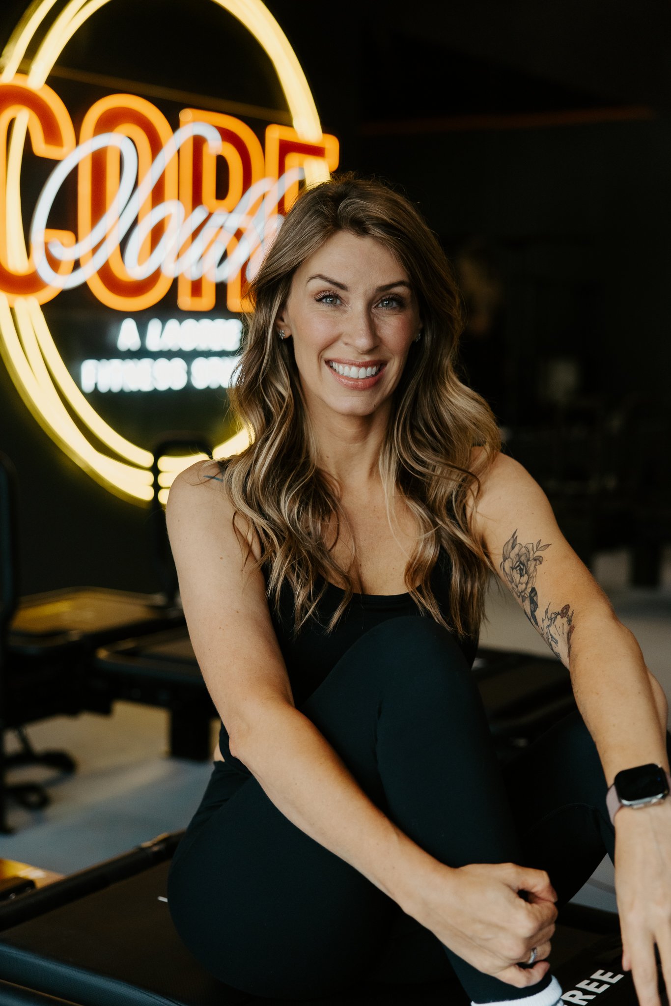 A smiling woman with long dark hair, wearing a black sleeveless top and black pants, stands with her hands on her hips in front of a neon sign that says 'Dough' and 'Dance Studio' in the background.