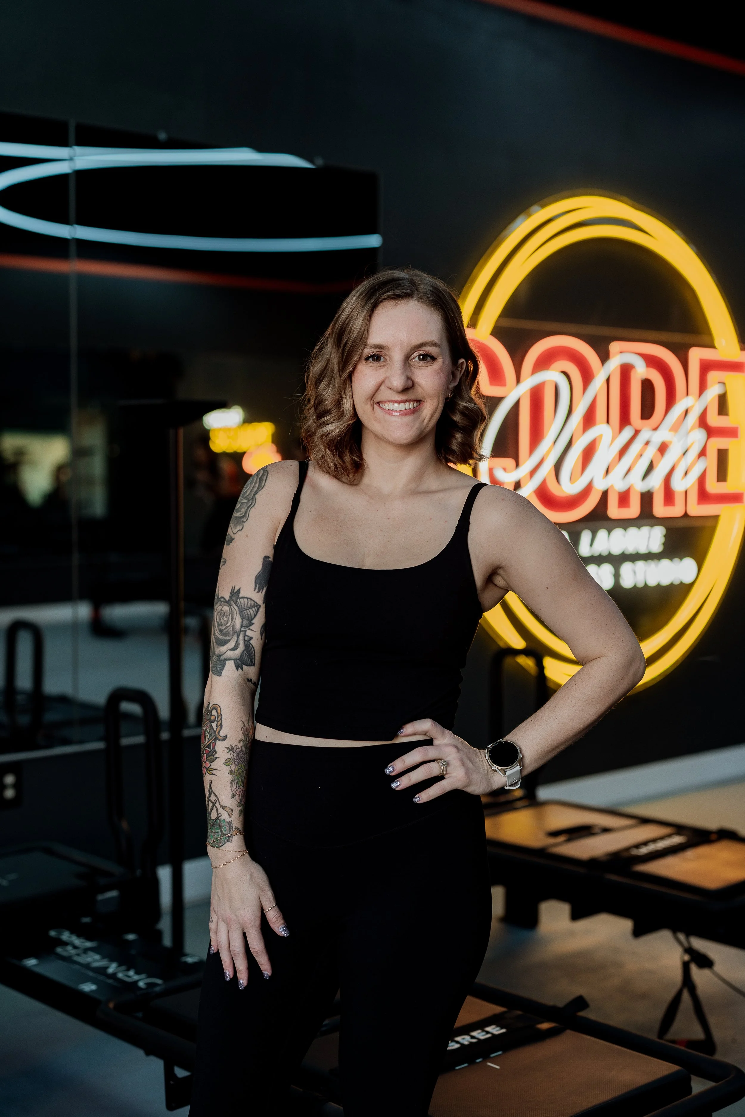 A woman with shoulder-length curly hair smiling, wearing a black sleeveless top and black pants, standing in front of neon signs inside a studio.