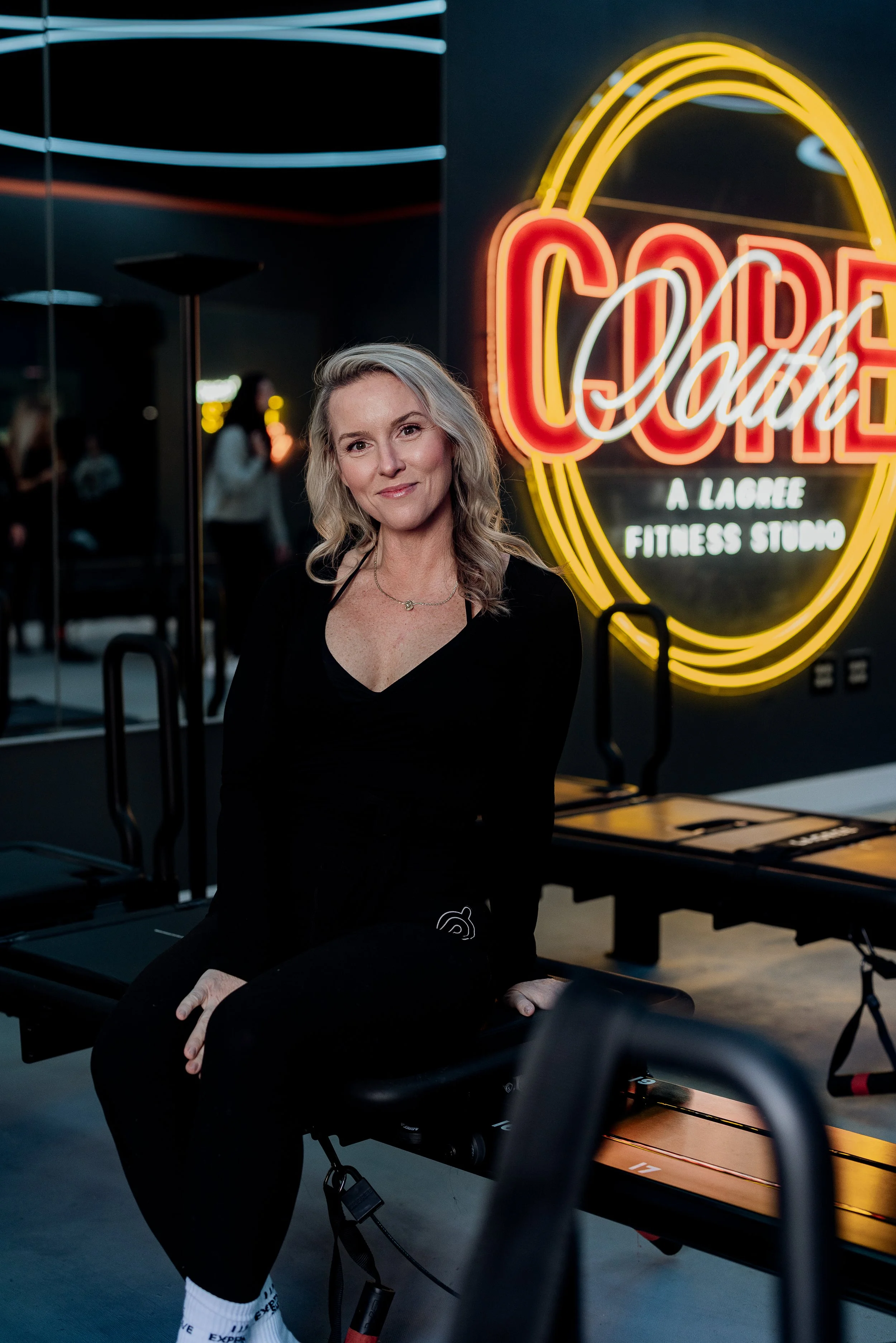 A woman with gray hair sitting on a reformer Pilates machine inside a fitness studio with a neon sign in the background reading "Code Your Fitness Studio."