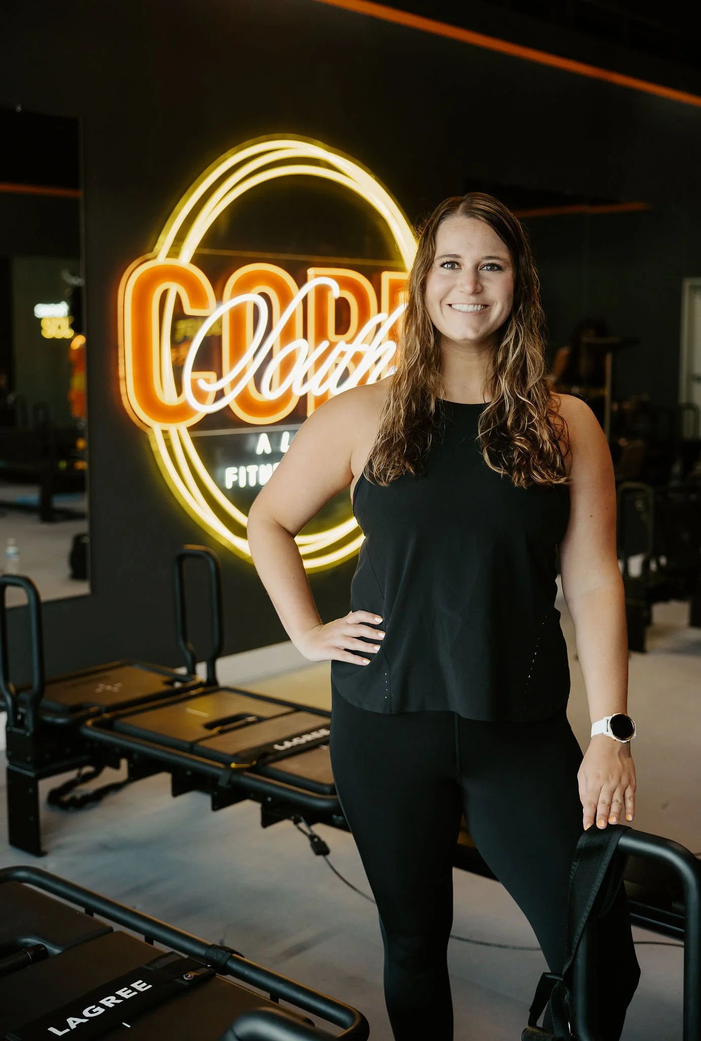 A smiling woman in black workout attire standing in a fitness studio with a neon sign that says 'Coach'. Exercise equipment is visible in the background.