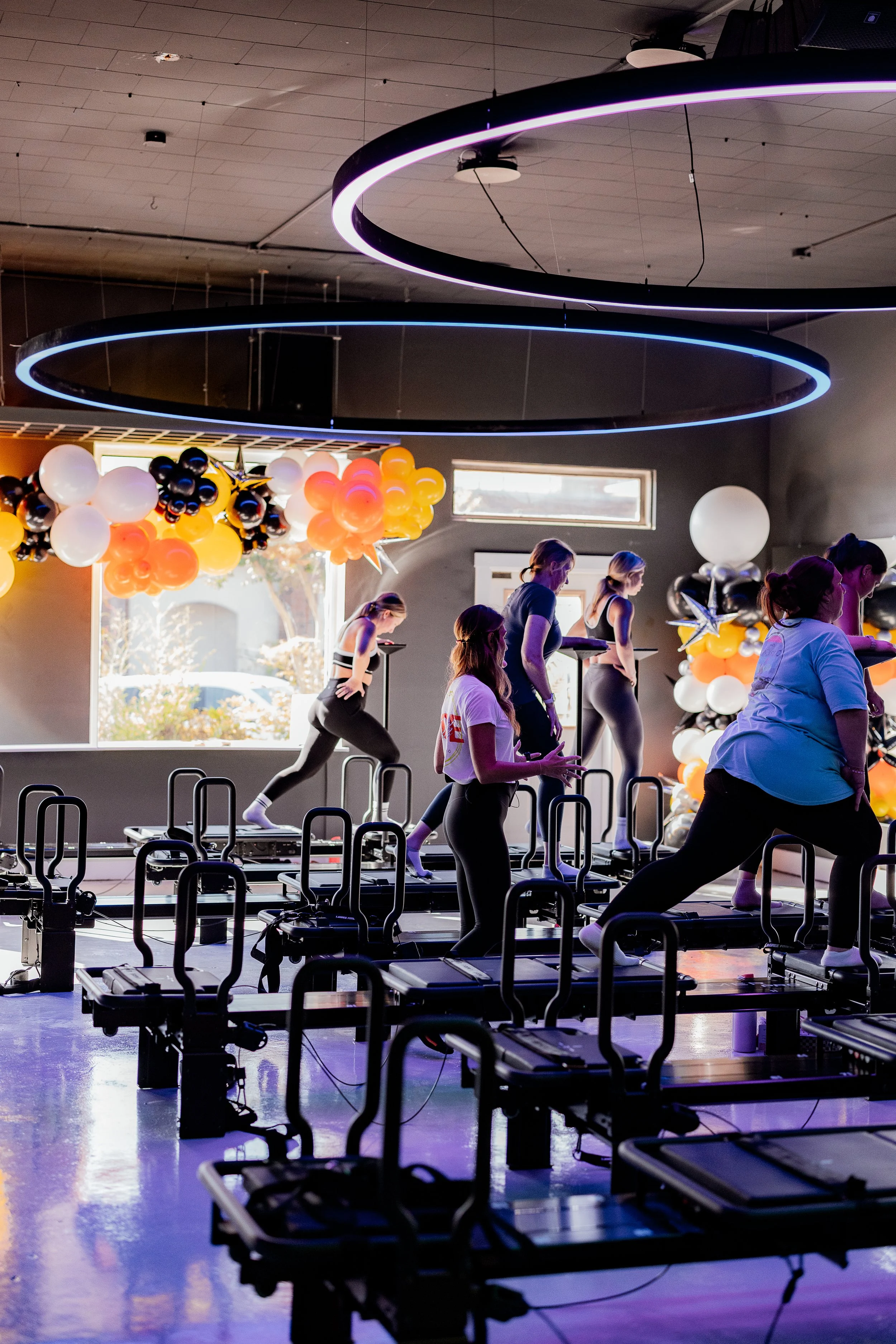 People participating in a fitness class indoors, using Lagree fitness machines, with colorful balloons and modern lighting fixtures hanging from the ceiling.