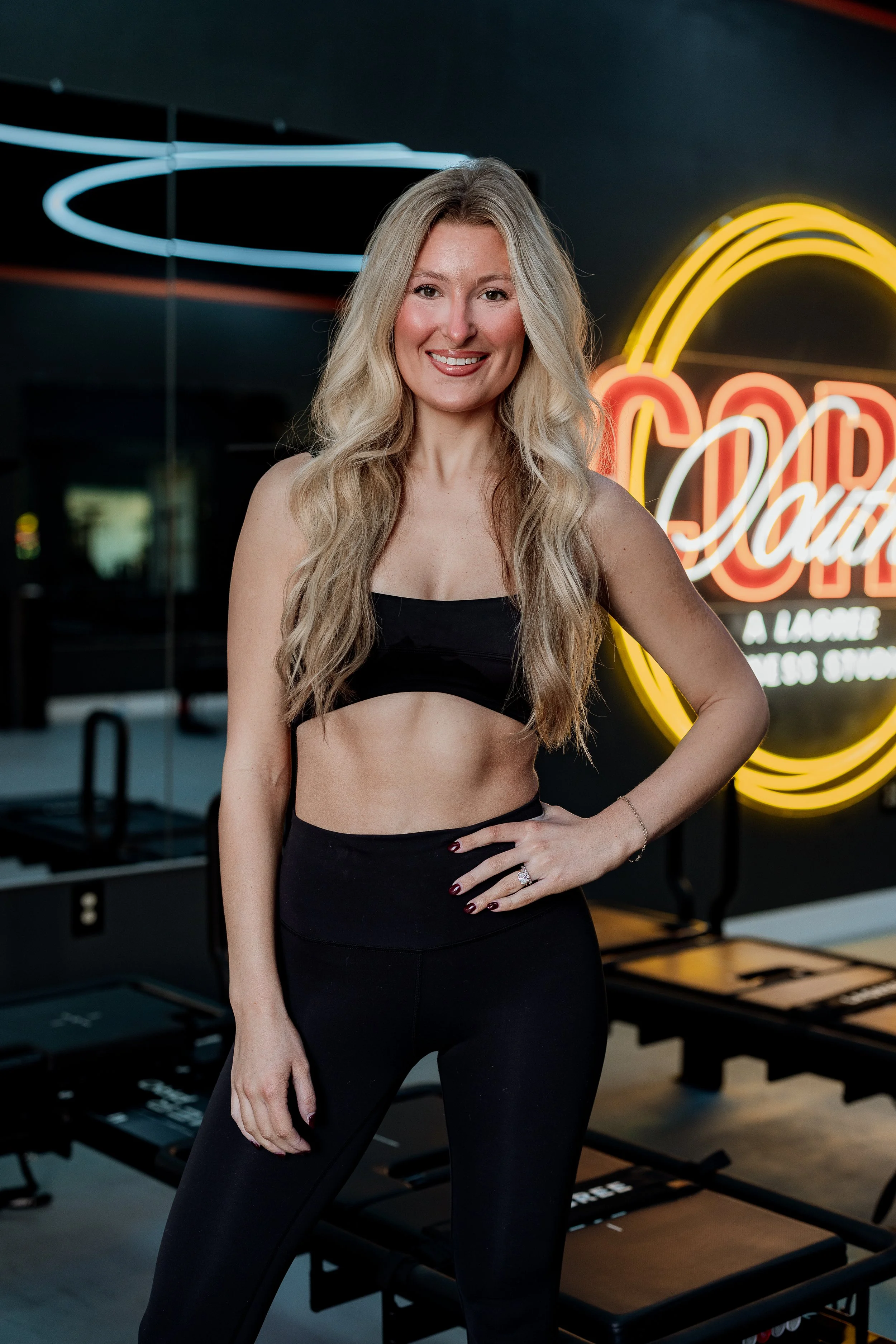 Woman with long blonde hair wearing a black sports bra and black leggings standing in front of a neon sign that says 'Cold' with a rainbow design. She is smiling with her right hand on her hip, in a fitness studio or gym.