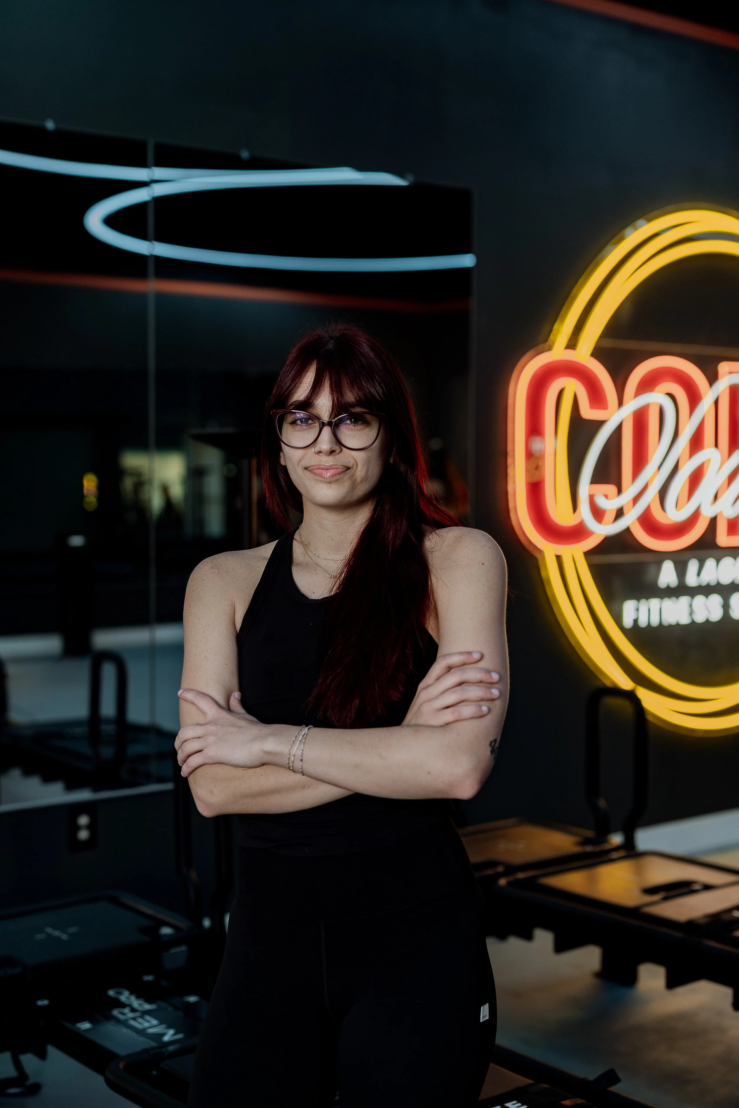 A young woman with long red hair, glasses, and a black sleeveless top stands with arms crossed in front of neon signs inside a fitness studio. The neon signs include a rainbow design and the word 'CO' in bright colors. The background shows exercise equipment and a dark wall.