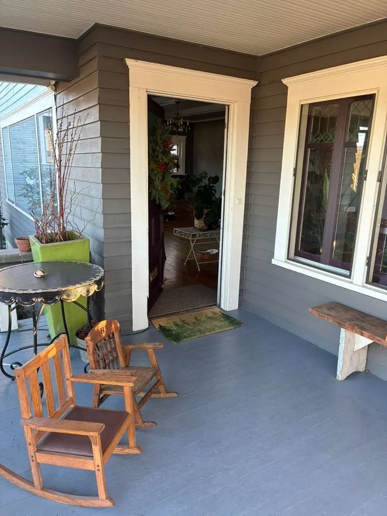 A porch with gray painted wooden floorboards and gray siding, featuring two small wooden rocking chairs, a small table with a decorative edge, and a window. The door to the house is open, revealing a room with plants and a Christmas tree inside.