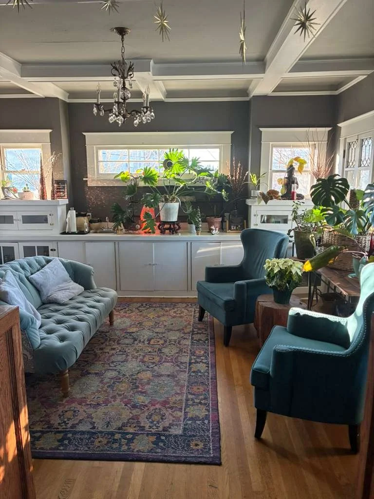 Living room with a teal tufted sofa, two matching teal wingback chairs, a floral area rug, and various potted plants on a white cabinet and side table. Sunlight streams through three windows, and a chandelier hangs from the ceiling.