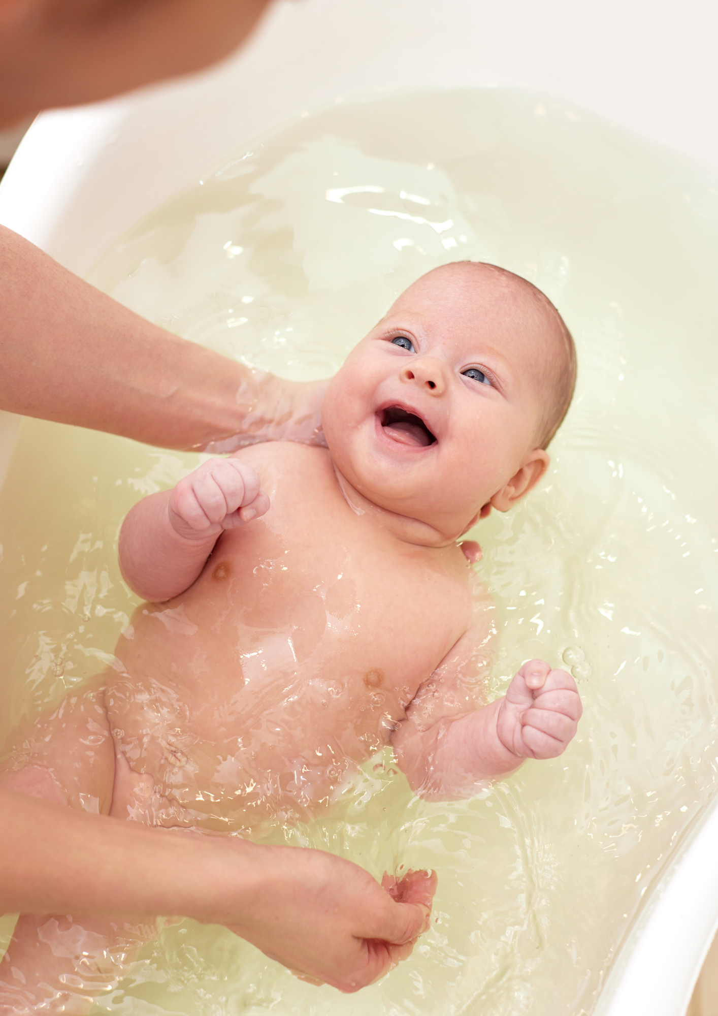 A baby being bathed in a white tub, smiling and looking up, with an adult supporting the baby.