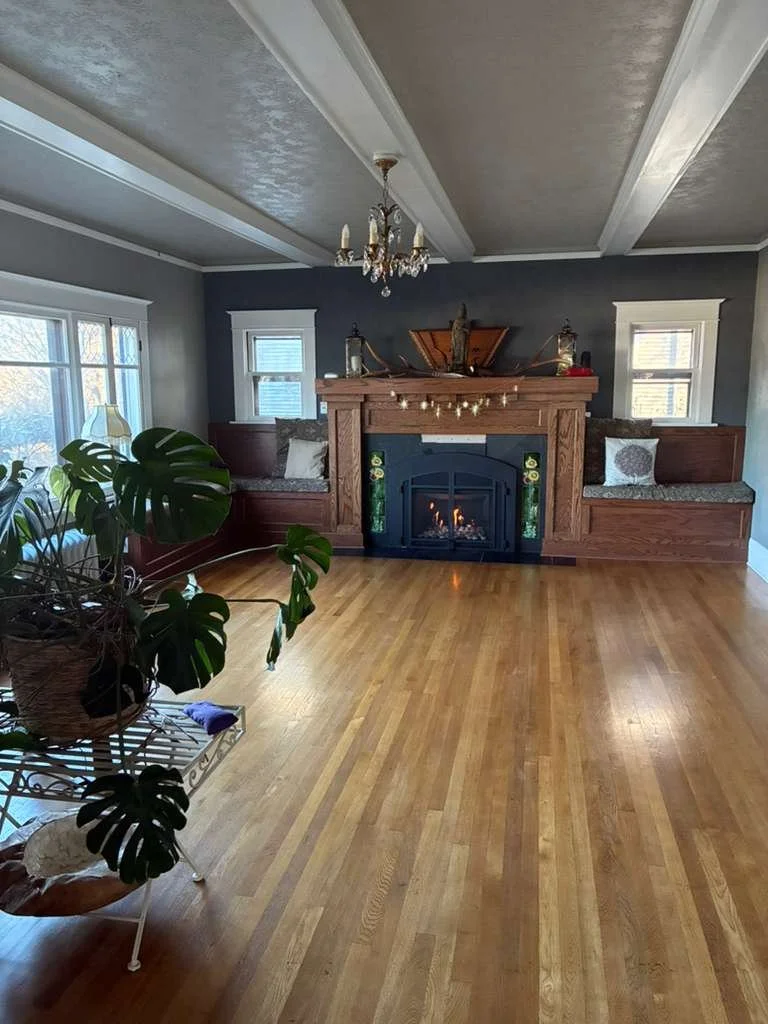 Living room with hardwood floor, gray walls, a fireplace with a wooden mantel, decorative items, a chandelier, and windows letting in natural light.