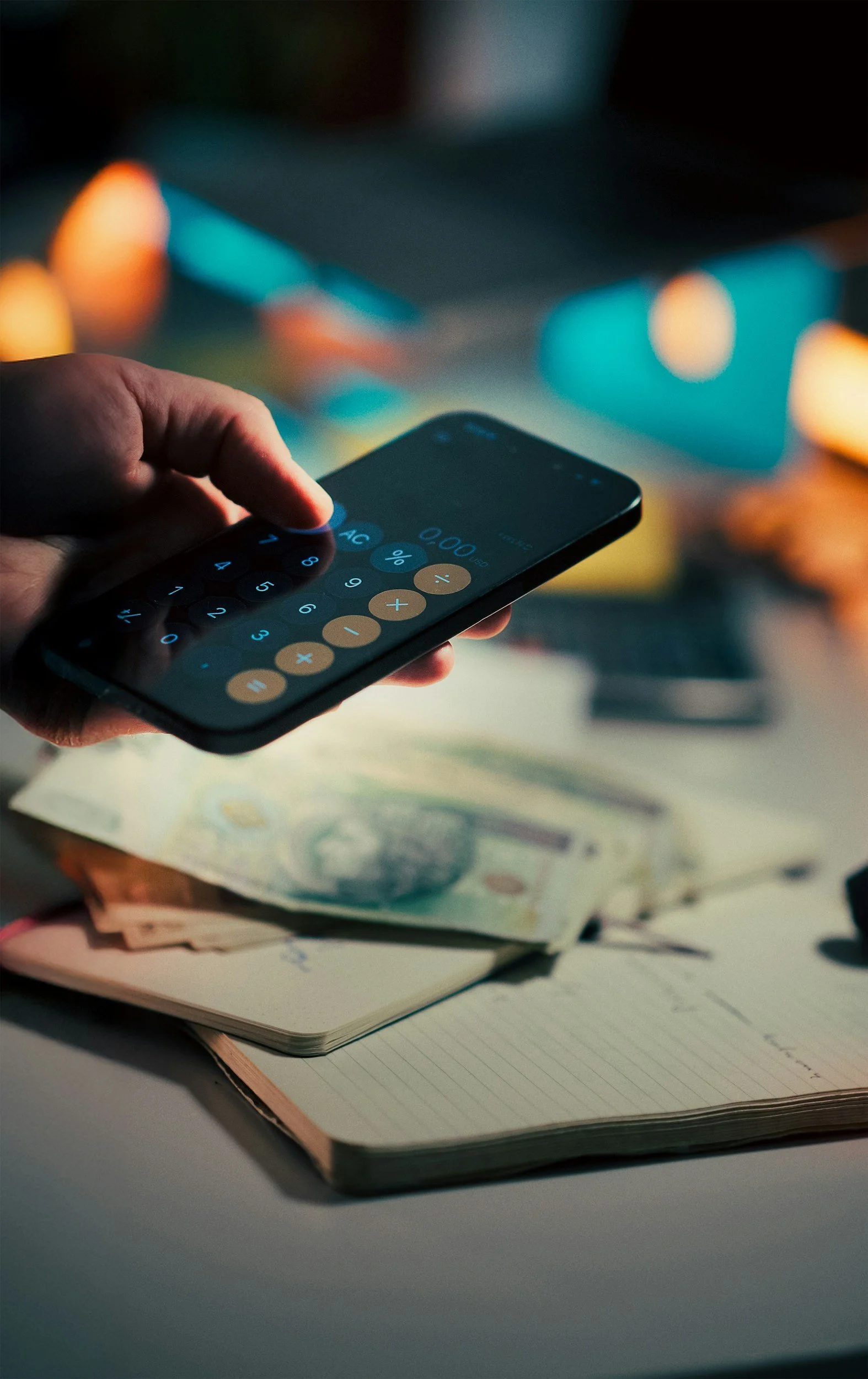 Hand holding a mobile phone with calculator app open, placed above a notebook and summer dollars bills on a desk.