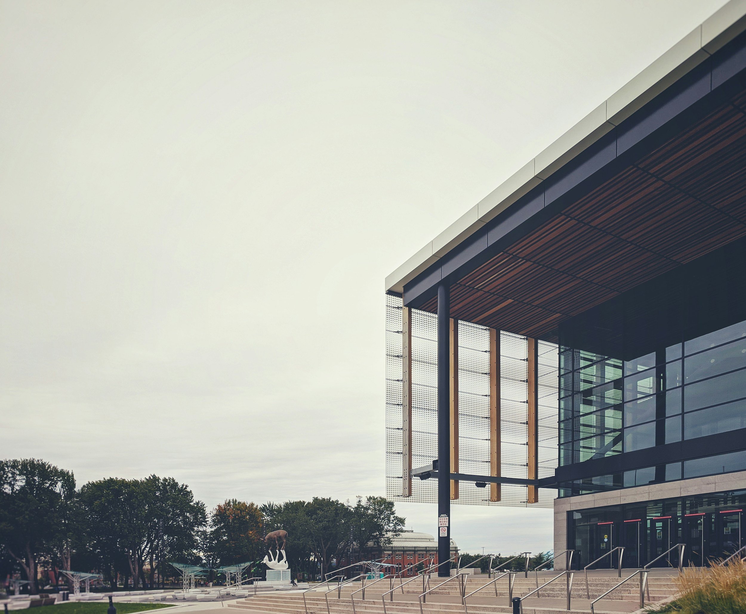 Modern building with glass and metal facade, trees and a sculpture of a swan in the background, overcast sky.