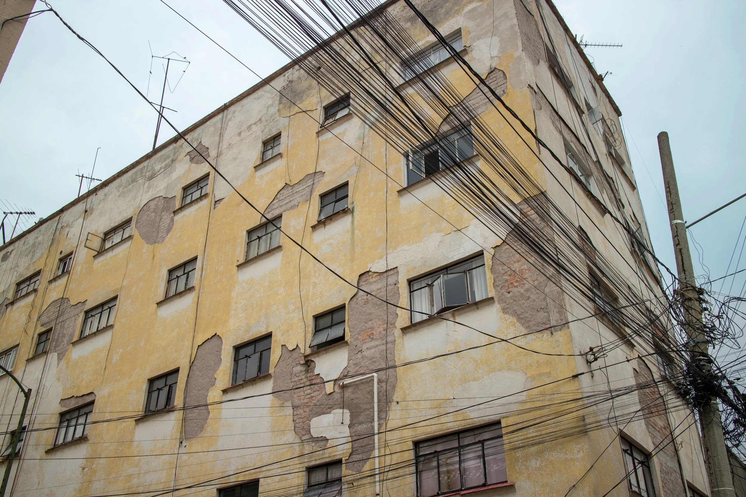 An old, multi-story building with yellow and peeling paint, broken windows, and exposed brickwork. Several electrical wires are crisscrossed in front of the building under an overcast sky.