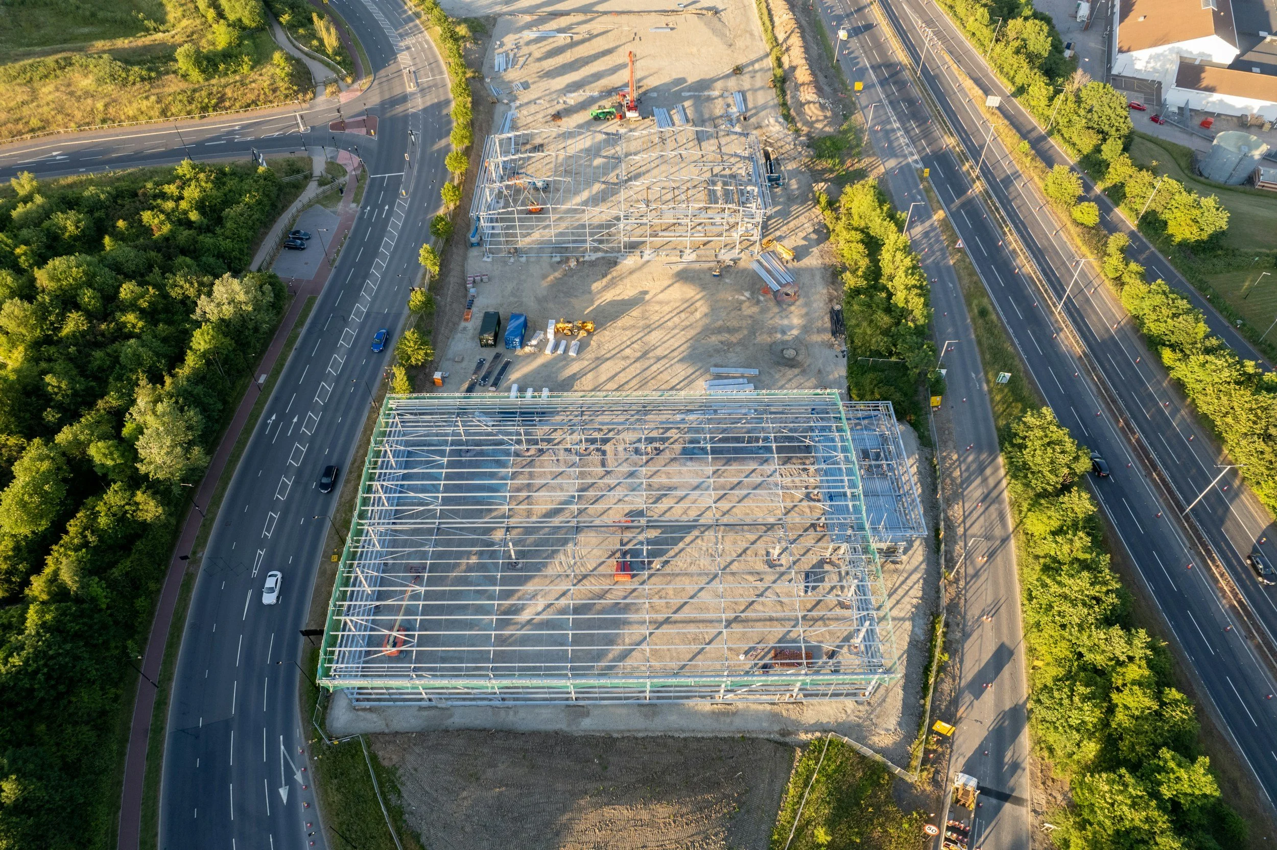 An aerial view of a construction site with metal framework being assembled for a building, surrounded by roads and lush green trees.