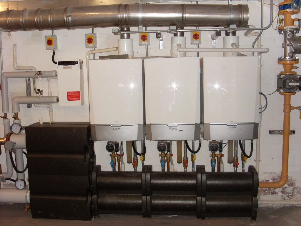 Three white boilers or heat exchangers connected to copper piping and industrial equipment, with black tanks below and control panels on the wall.