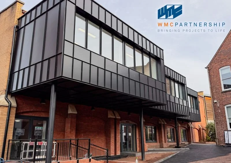 Modern brick building with black metal-framed glass extension on the second floor, supported by black columns. WMC Partnership logo and slogan "Bringing Projects to Life" visible at the top right.