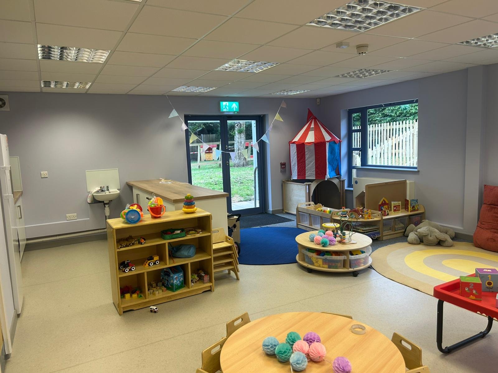 Indoor children’s playroom and activity area with toys, furniture, and decorations, including a circus tent, bunting, small tables, and a play area with a carpet.