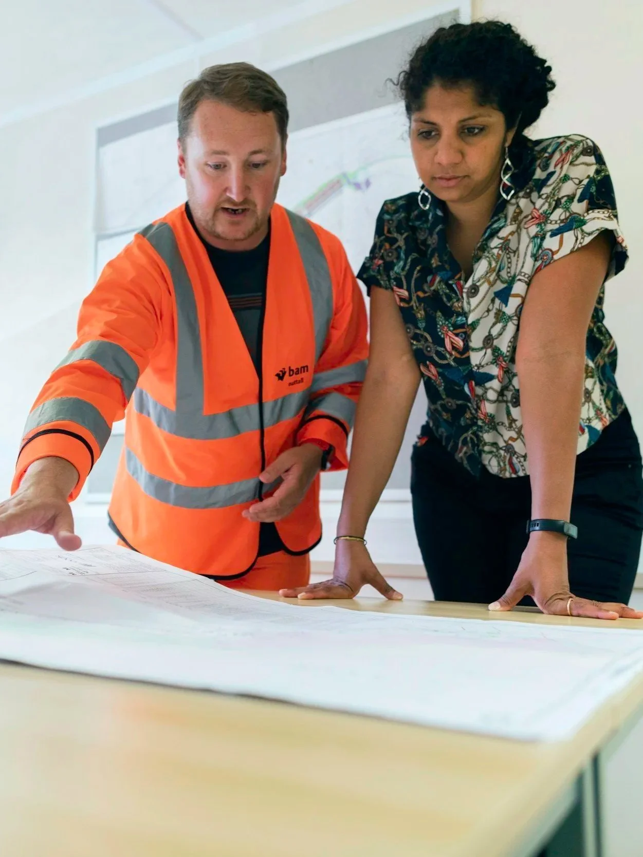 Two professionals, one in an orange high-visibility vest and the other in a patterned blouse, examine large architectural blueprints on a table in a bright office.
