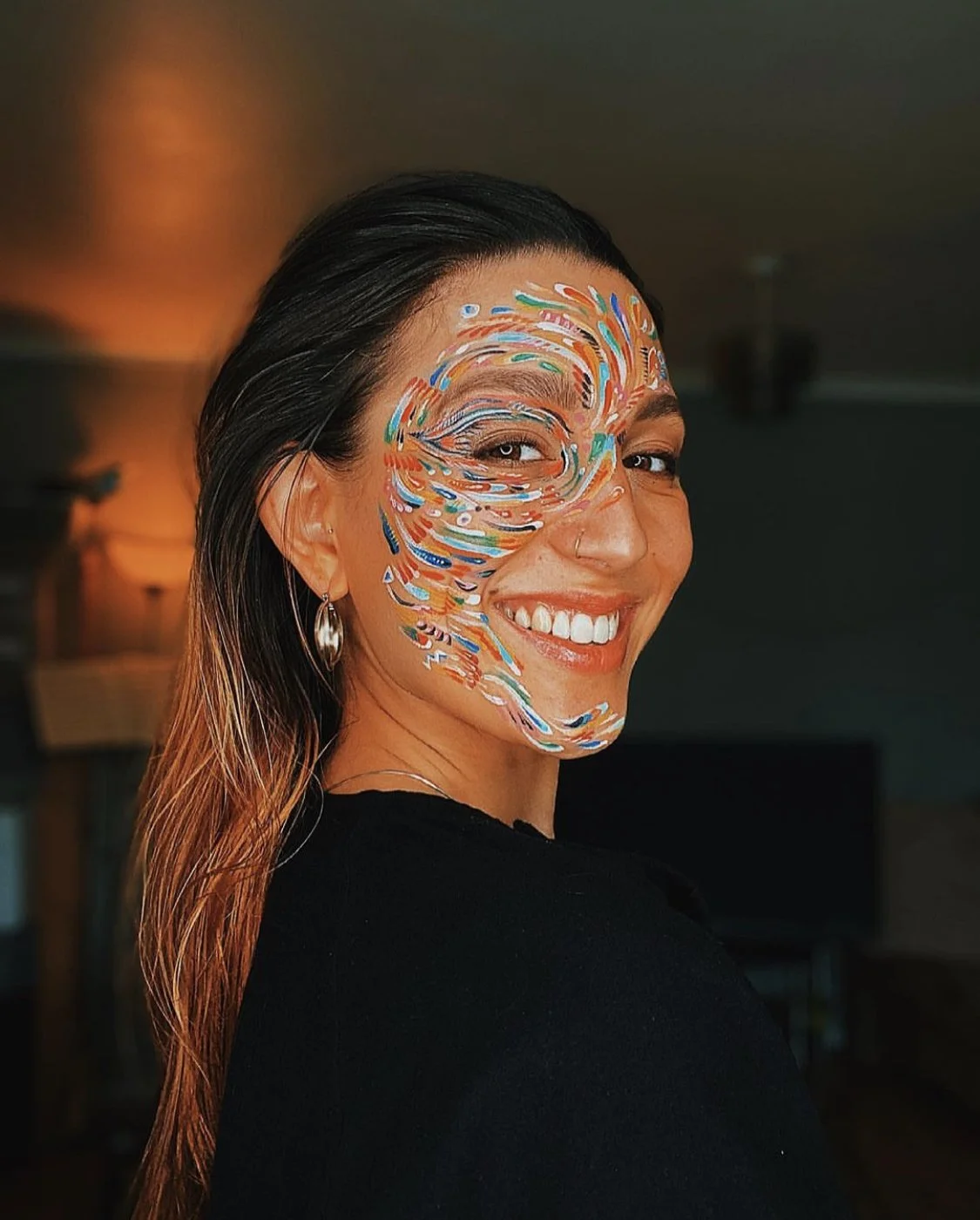 Ning-Ning is with long hair, earrings, and a black top, smiling with colourful face paint on the left side of her face in an indoor setting.