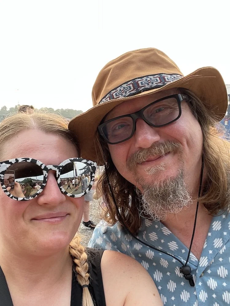 A woman with blonde hair in a braid wearing large zebra-patterned sunglasses, and a man with long hair, glasses, a beard and mustache, wearing a wide-brimmed hat and a patterned shirt, taking a selfie at an outdoor event.