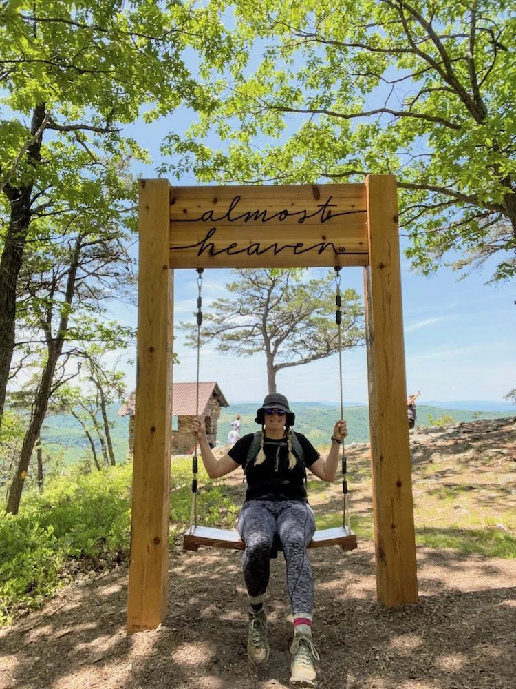 Woman sitting on a swing under a wooden frame with the sign 'almost heaven' in a scenic outdoor area with trees, greenery, and a mountain view.