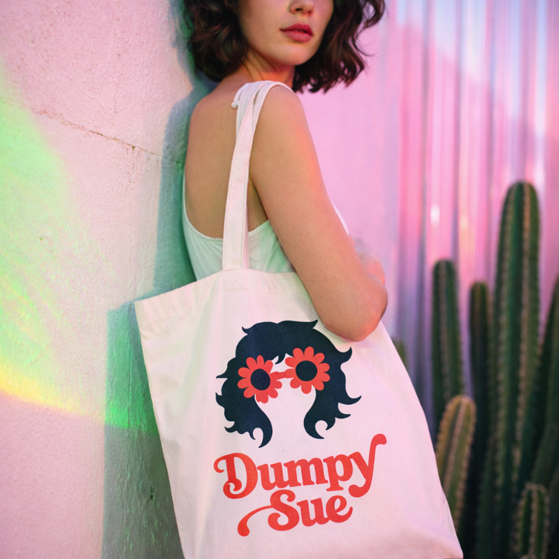 A woman with curly dark hair wearing a white tank top, holding a tote bag with a graphic of a woman with glasses and flowers in her hair, and the words "Dumpy Sue" printed on it, standing near a cactus plant against a colorful, neon-lit background.