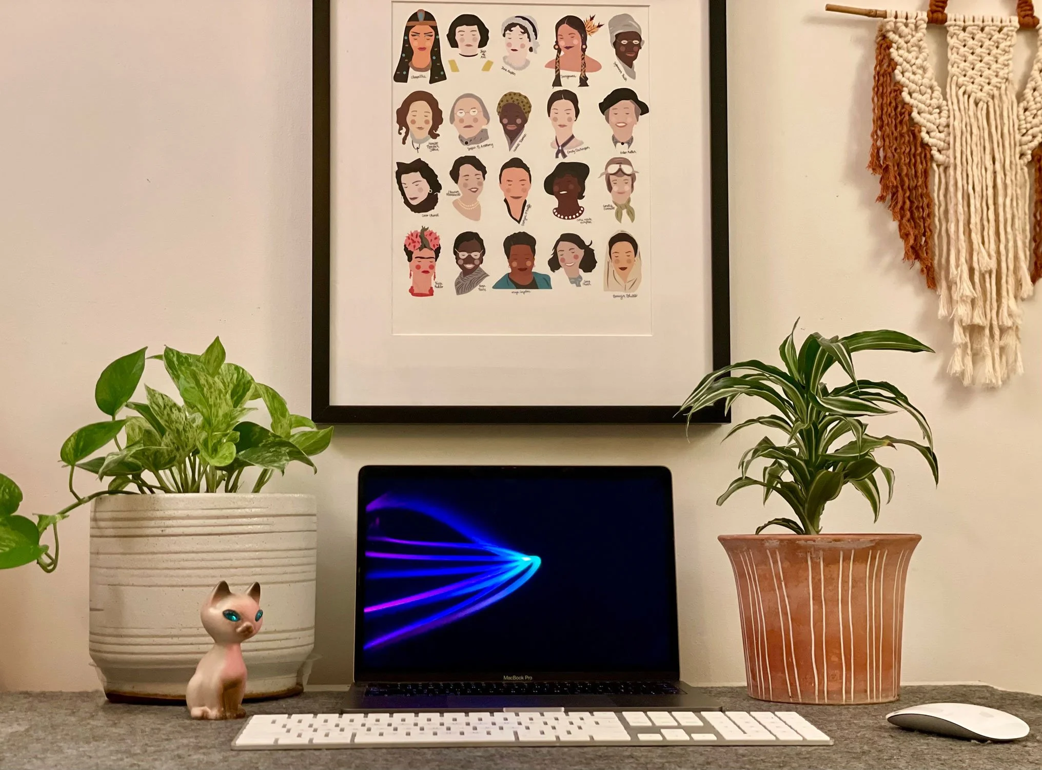 A desk setup with two potted plants, a MacBook Pro, a white keyboard, and a white mouse. Behind the laptop, there is a framed illustration of diverse women and a woven wall hanging.