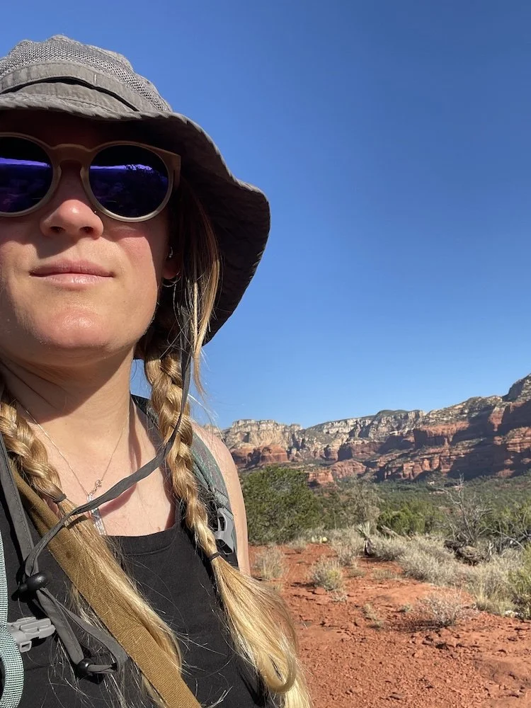 A woman with blonde braids wearing sunglasses and a wide-brimmed hat, hiking in a desert landscape with red dirt and red rock formations in the background.