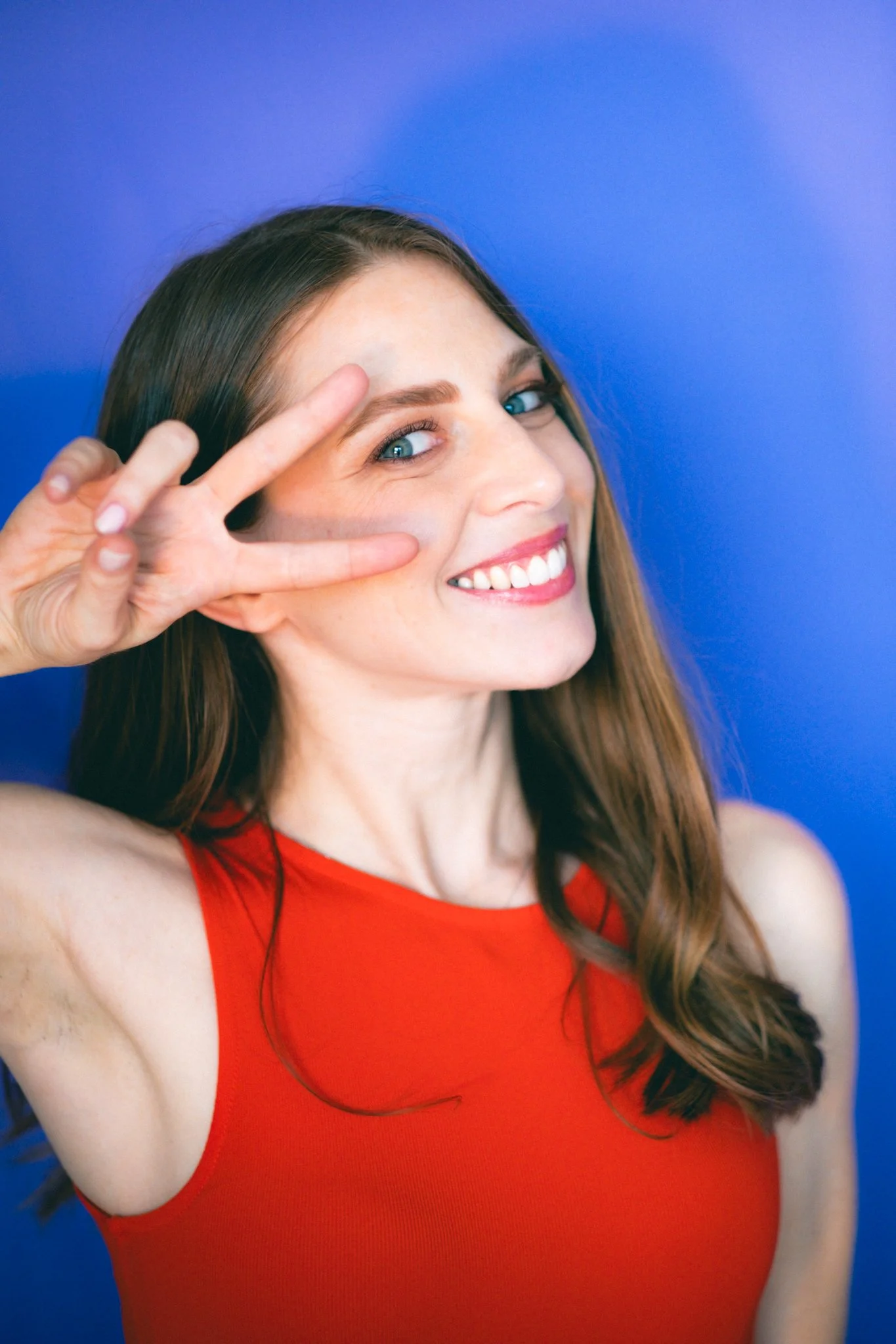 Una donna sorridente con capelli lunghi castani, occhi azzurri, che indossa un top rosso, fa un gesto di pace con due dita vicino all'occhio, contro uno sfondo blu.