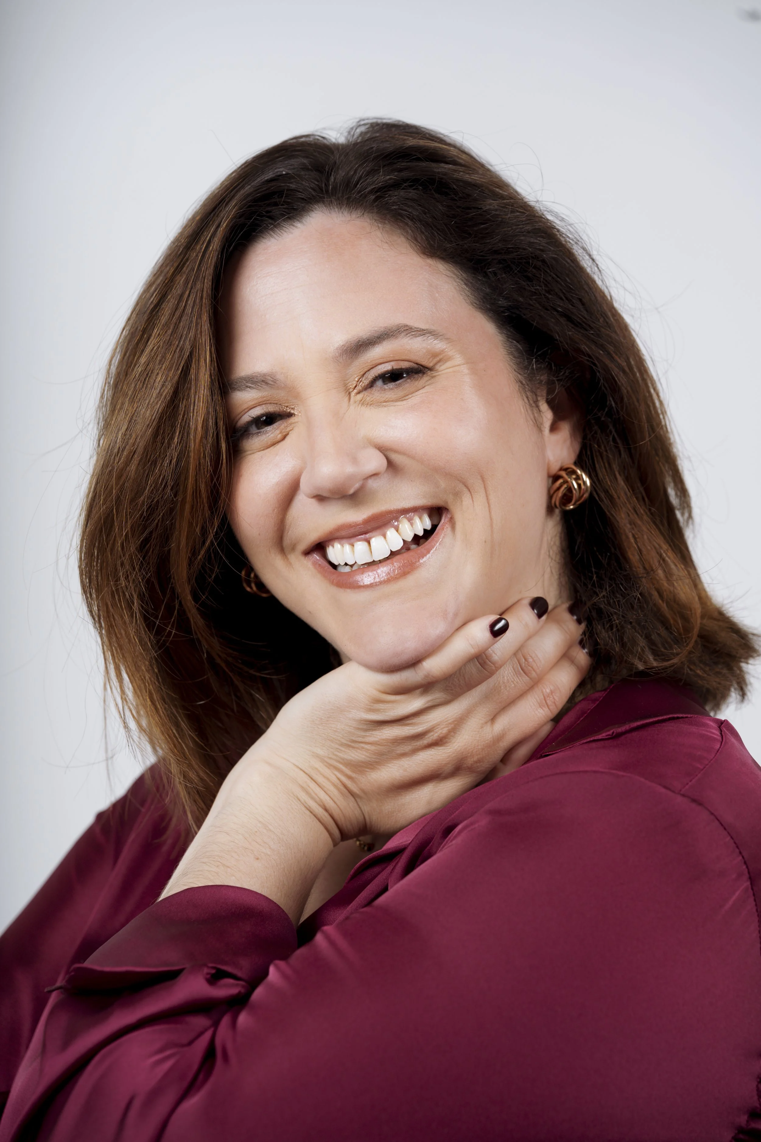 Una donna sorridente con capelli castani, indossa un top bordeaux, con orecchini rotondi d'oro, attira attenzione con espressione felice e posa con la mano sotto il mento.