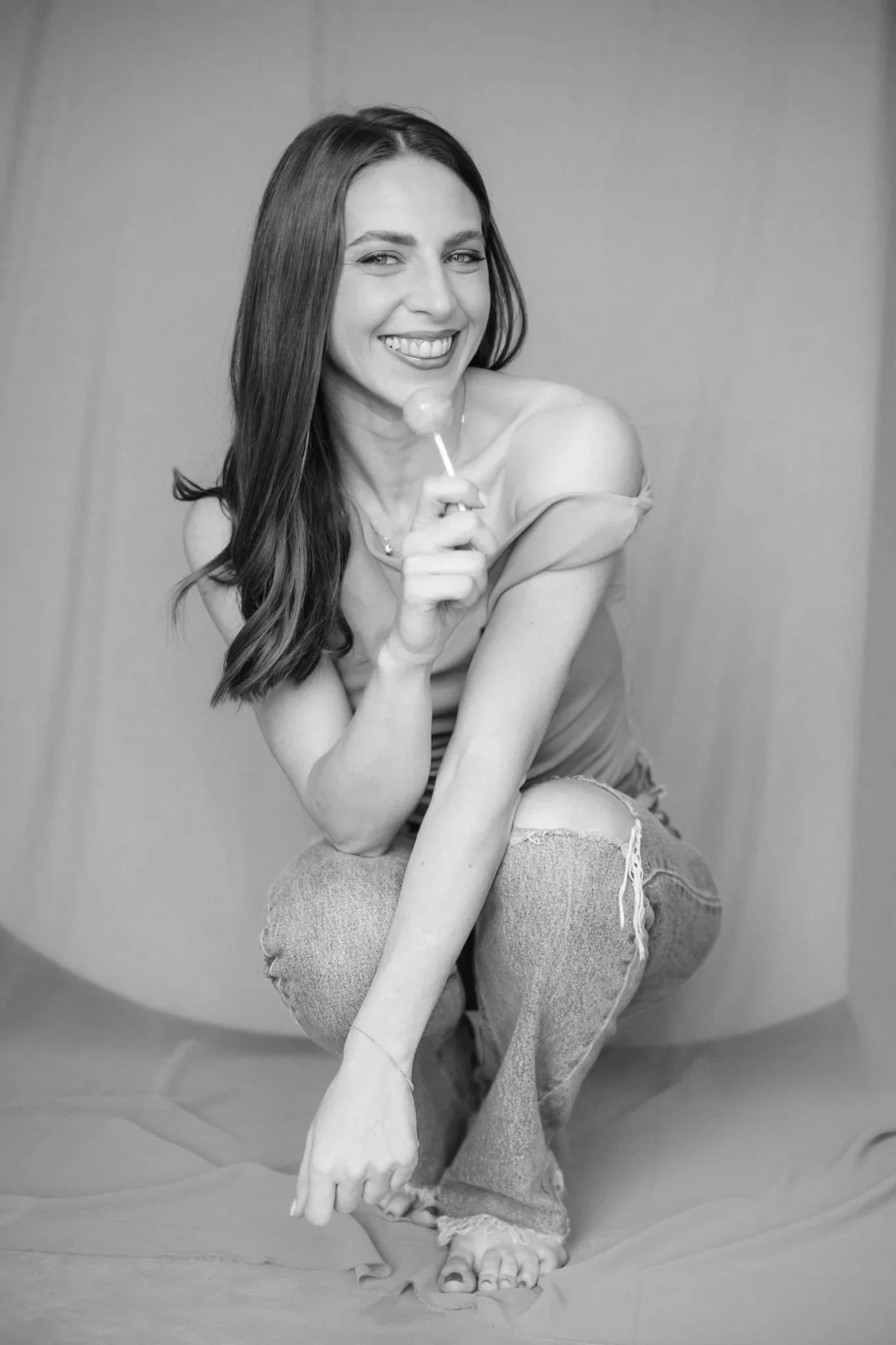 Una giovane donna con lunghi capelli castani, sorridente, seduta su un pavimento, che tiene un lecca lecca, abbigliamento casual con jeans strappati, in bianco e nero.