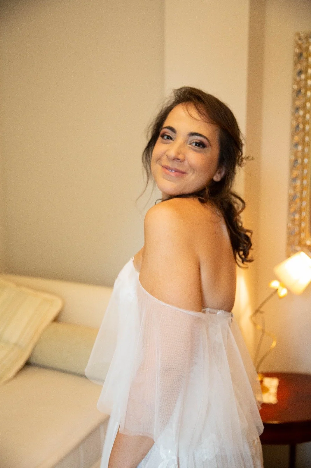 Una donna sorridente con capelli castani riccioli, indossa un abito bianco semi trasparente e sembra essere in un ambiente interno ben illuminato.