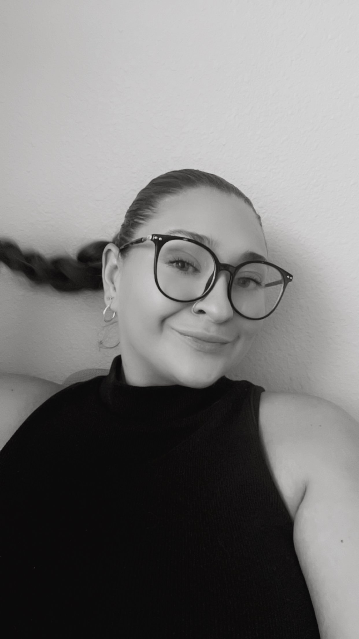 A young woman with glasses, earrings, and a nose ring smiling at the camera, with her hair pulled back and a braid sticking out behind her head, against a plain wall.