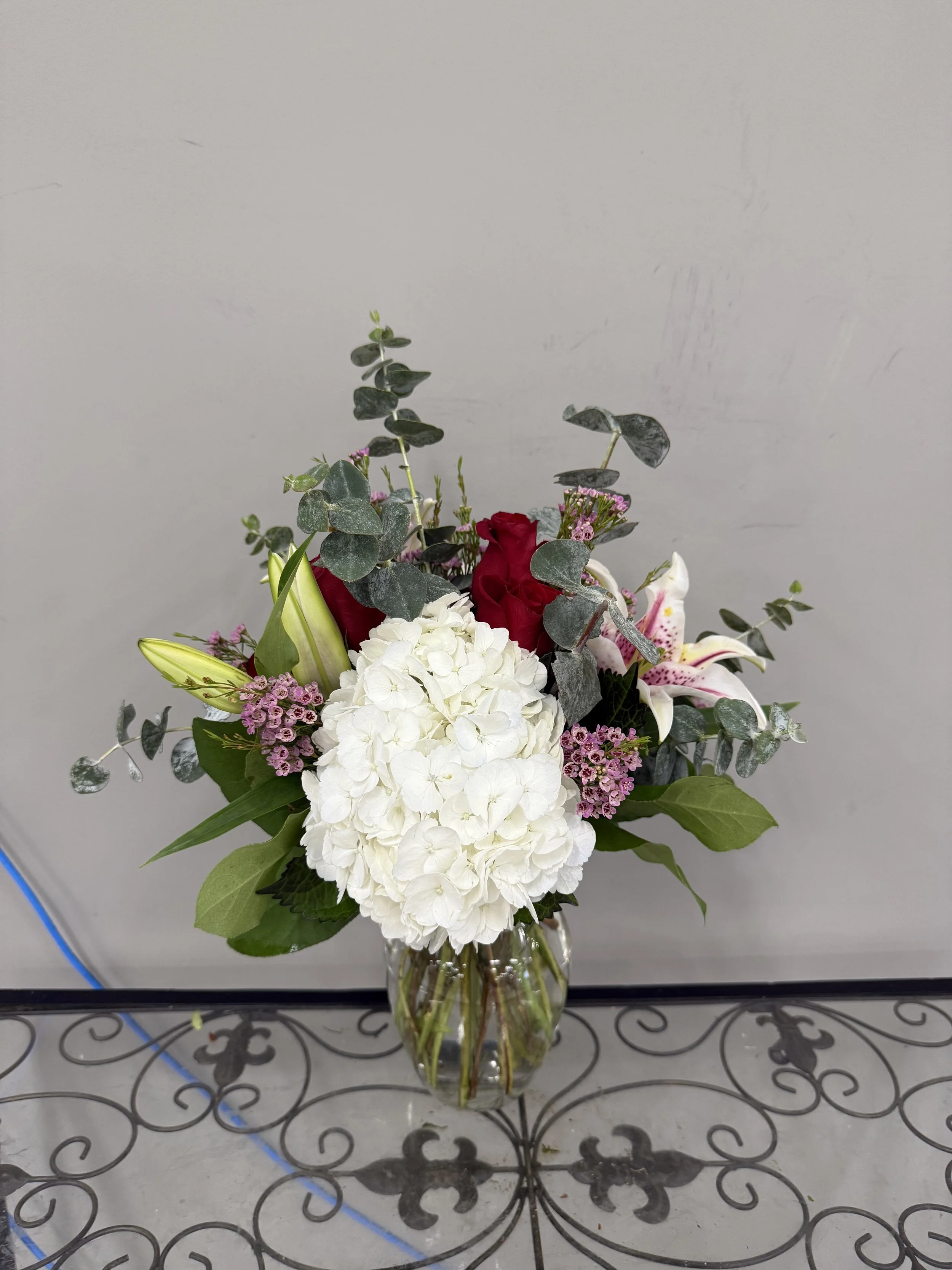 A bouquet of flowers including white hydrangeas, red roses, pink lilies, and purple small filler flowers in a clear glass vase, set on a decorative metal table against a plain wall.